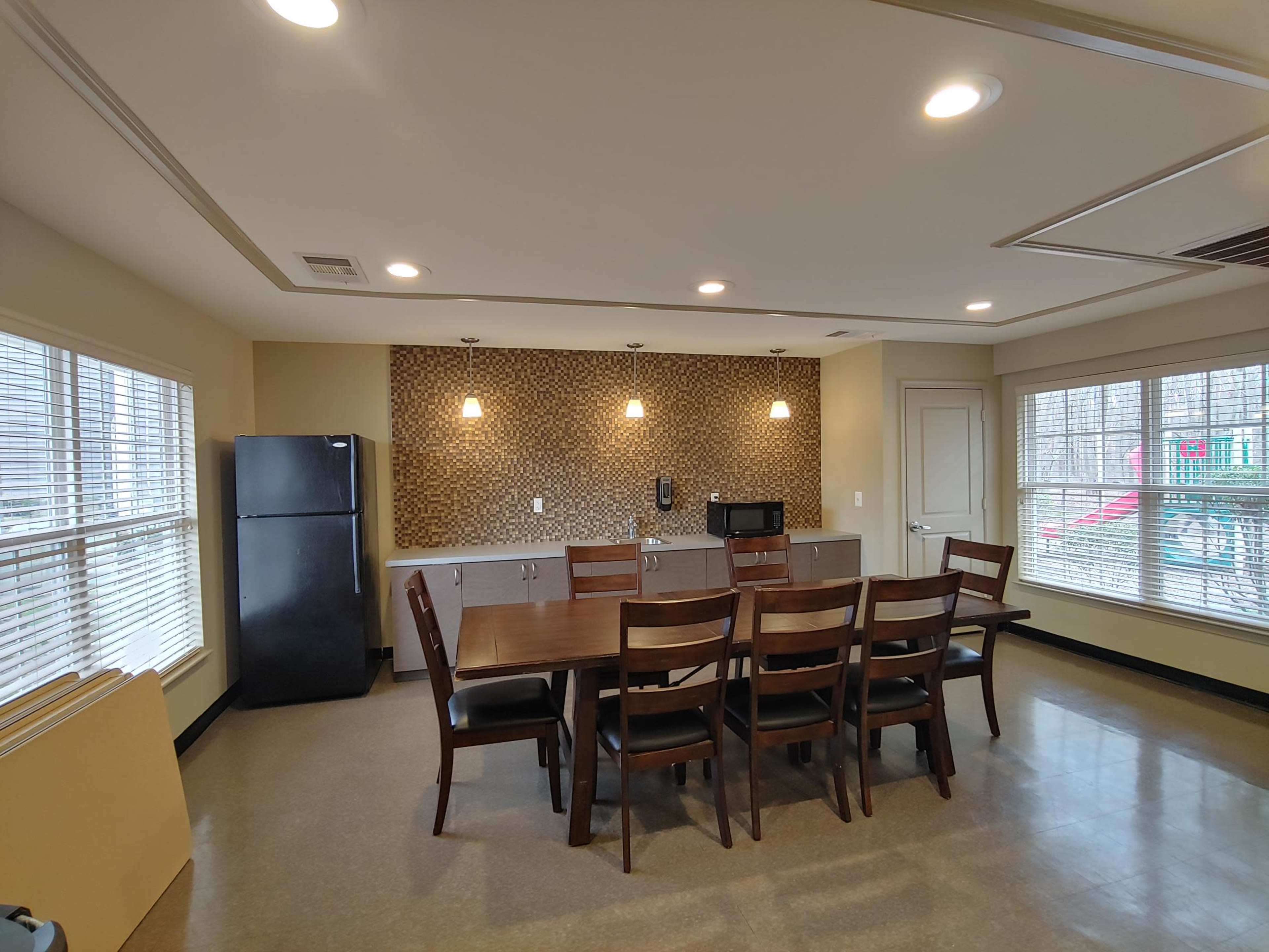 A spacious dining area features a wooden table with eight chairs, a refrigerator, a microwave, and large windows allowing natural light to enter.