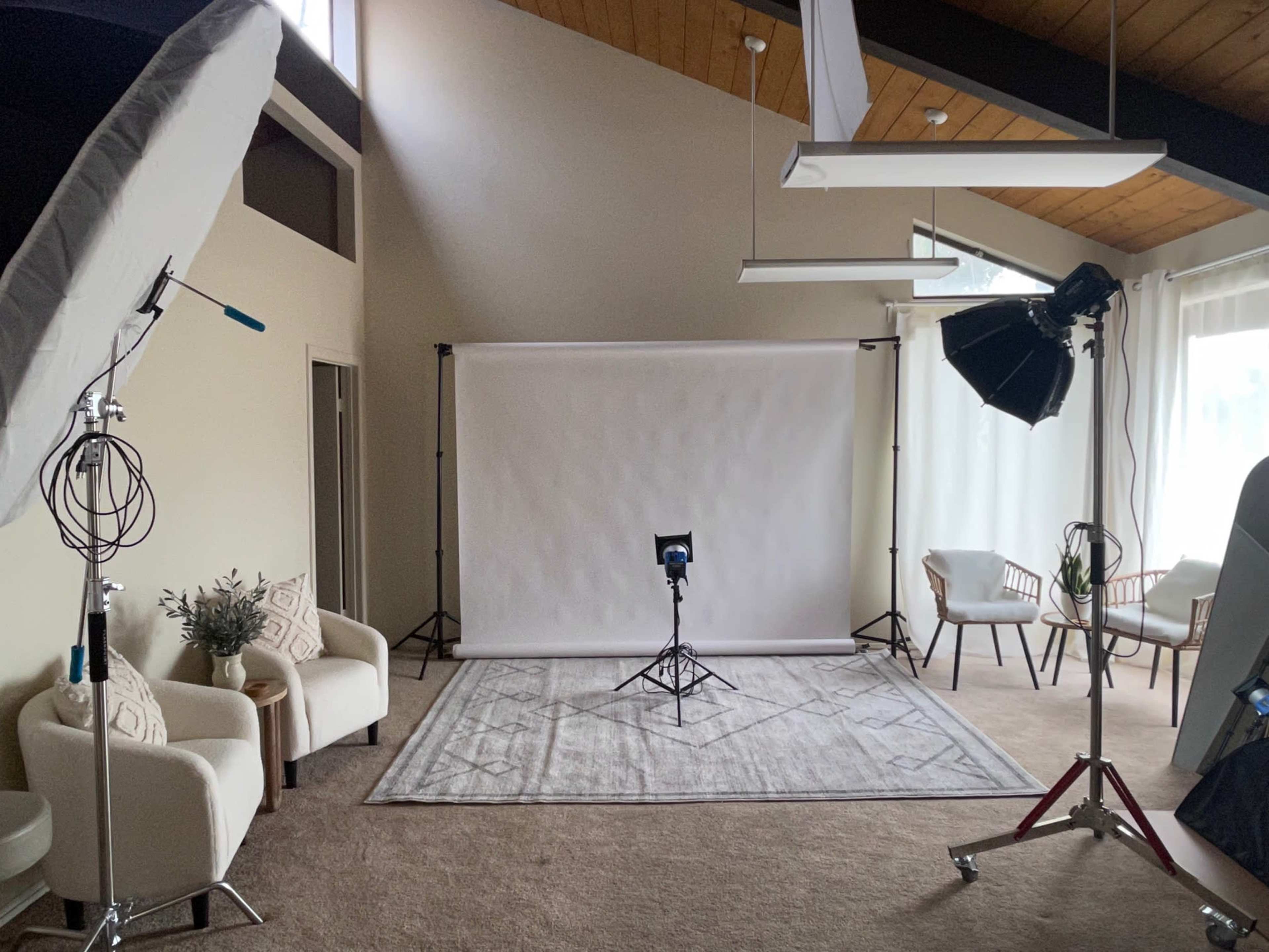 The image shows a studio setup with lighting equipment, a backdrop, and seating in a spacious room with high ceilings and large windows.