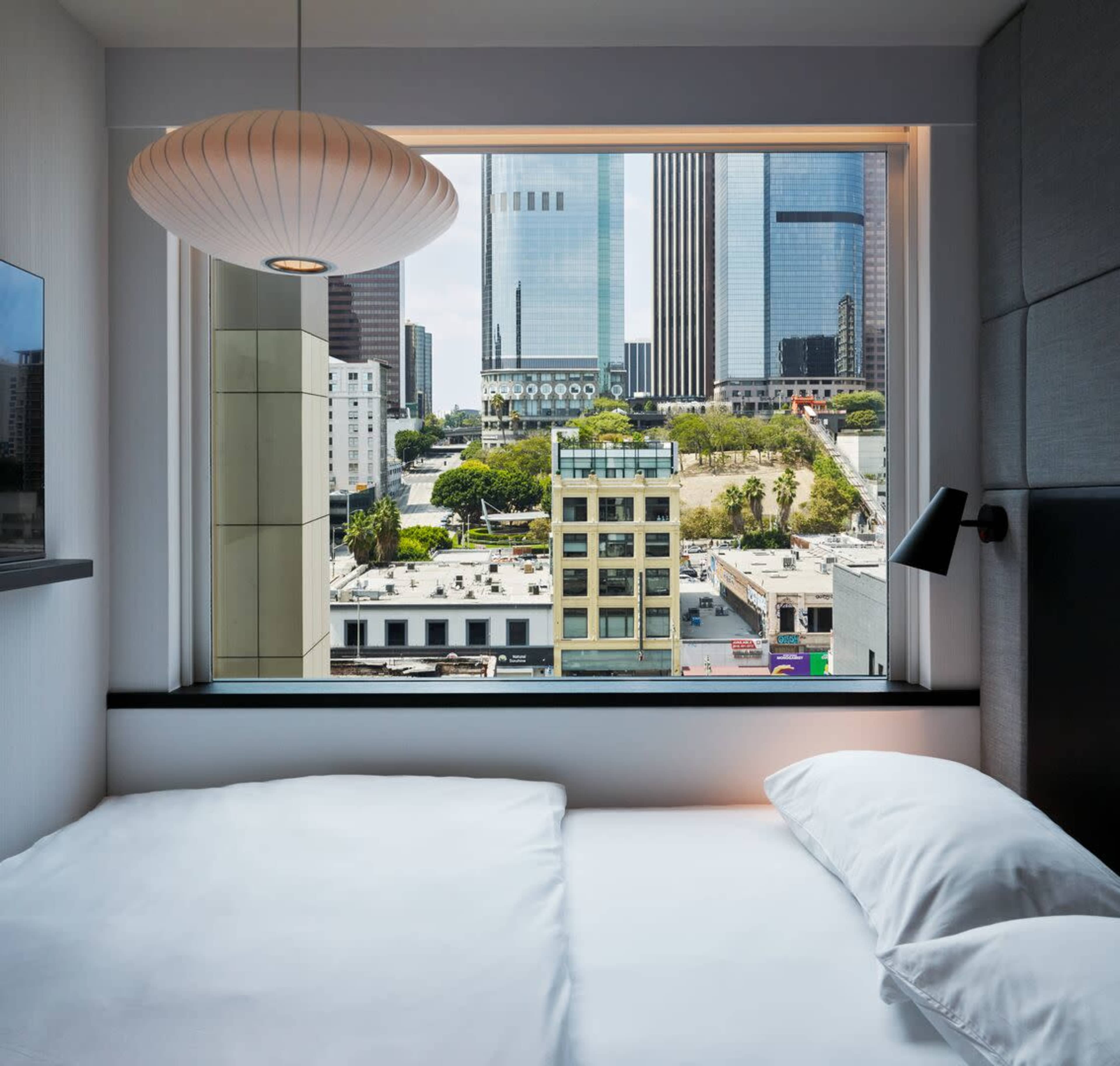 The image shows a hotel room with a neatly made bed and a large window offering a view of a cityscape with modern buildings and greenery.