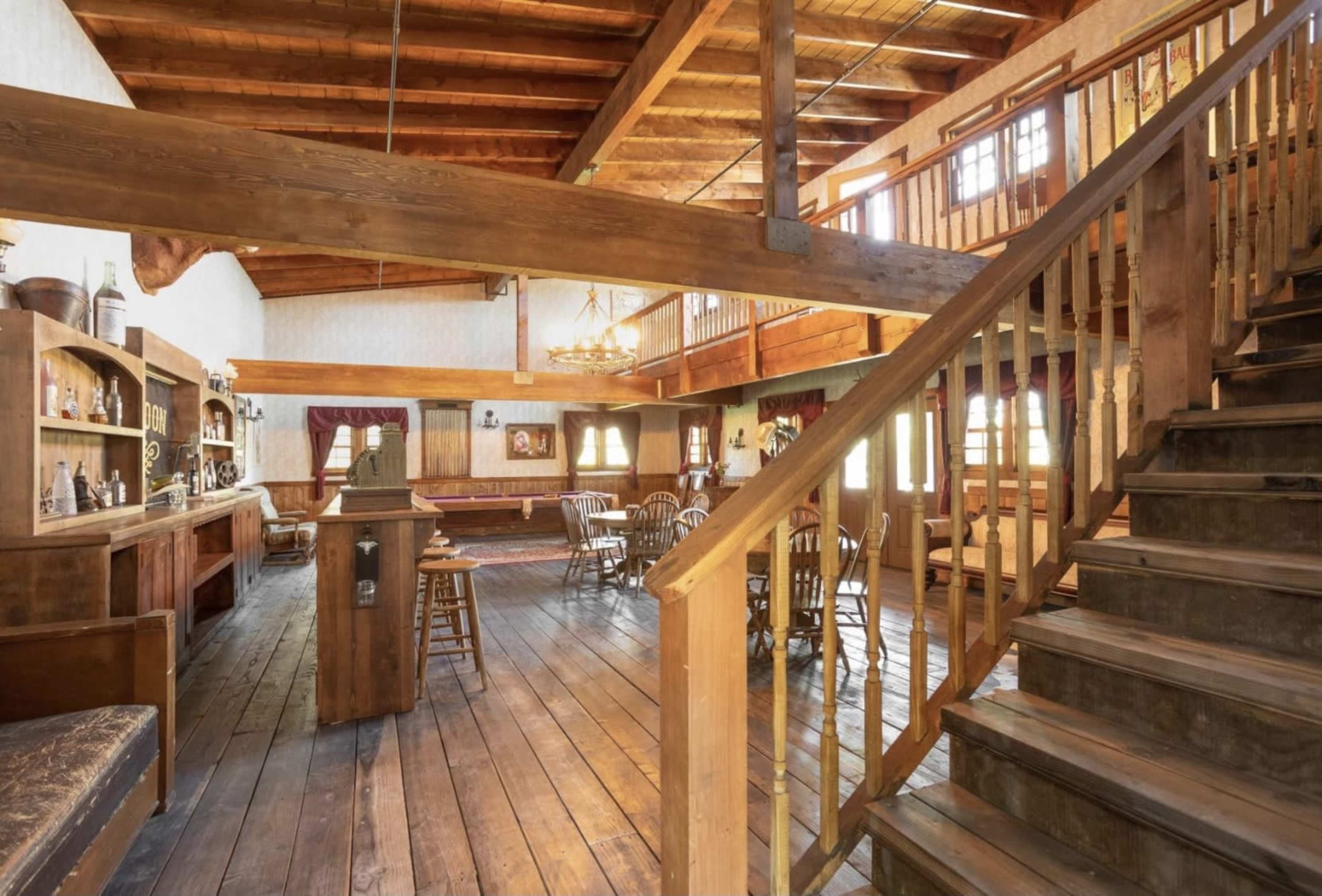 The interior of a rustic space featuring wooden beams, a staircase leading to a loft, and a bar area with seating.