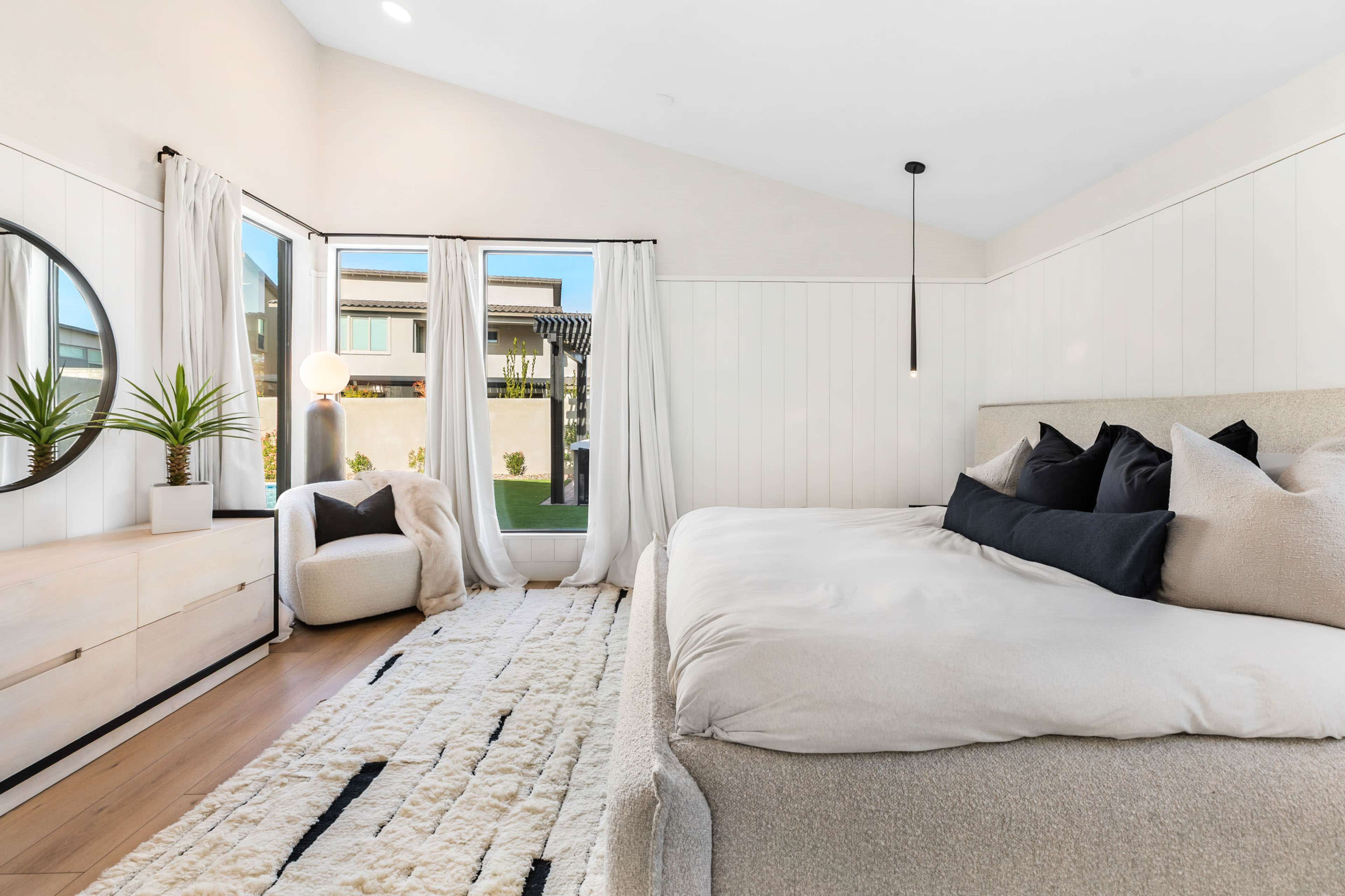 The image shows a modern bedroom with a bed, a round mirror, a cozy seating area, and large windows letting in natural light.