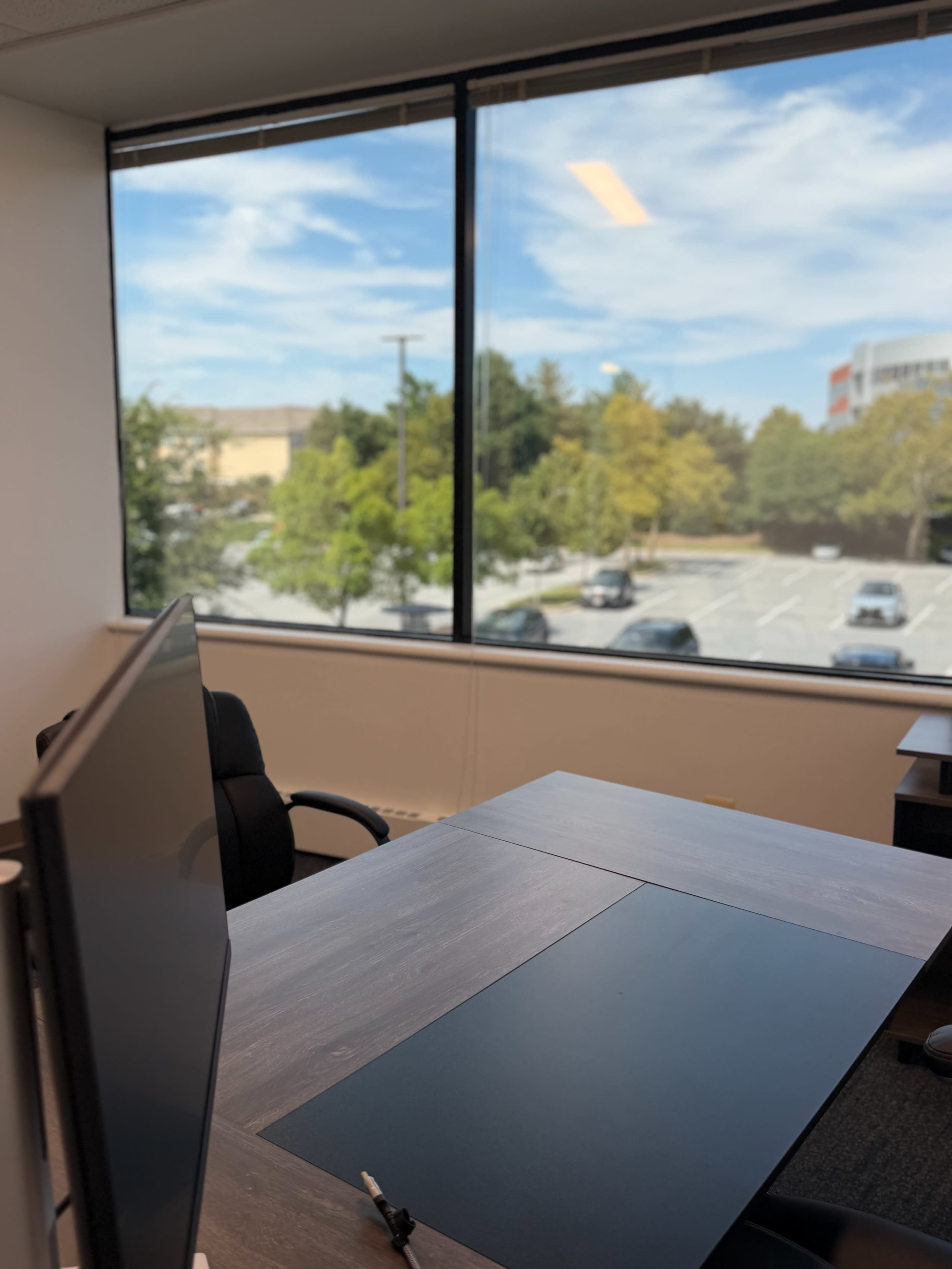 An office space features a desk and a computer monitor, with a large window overlooking a parking lot and trees outside.