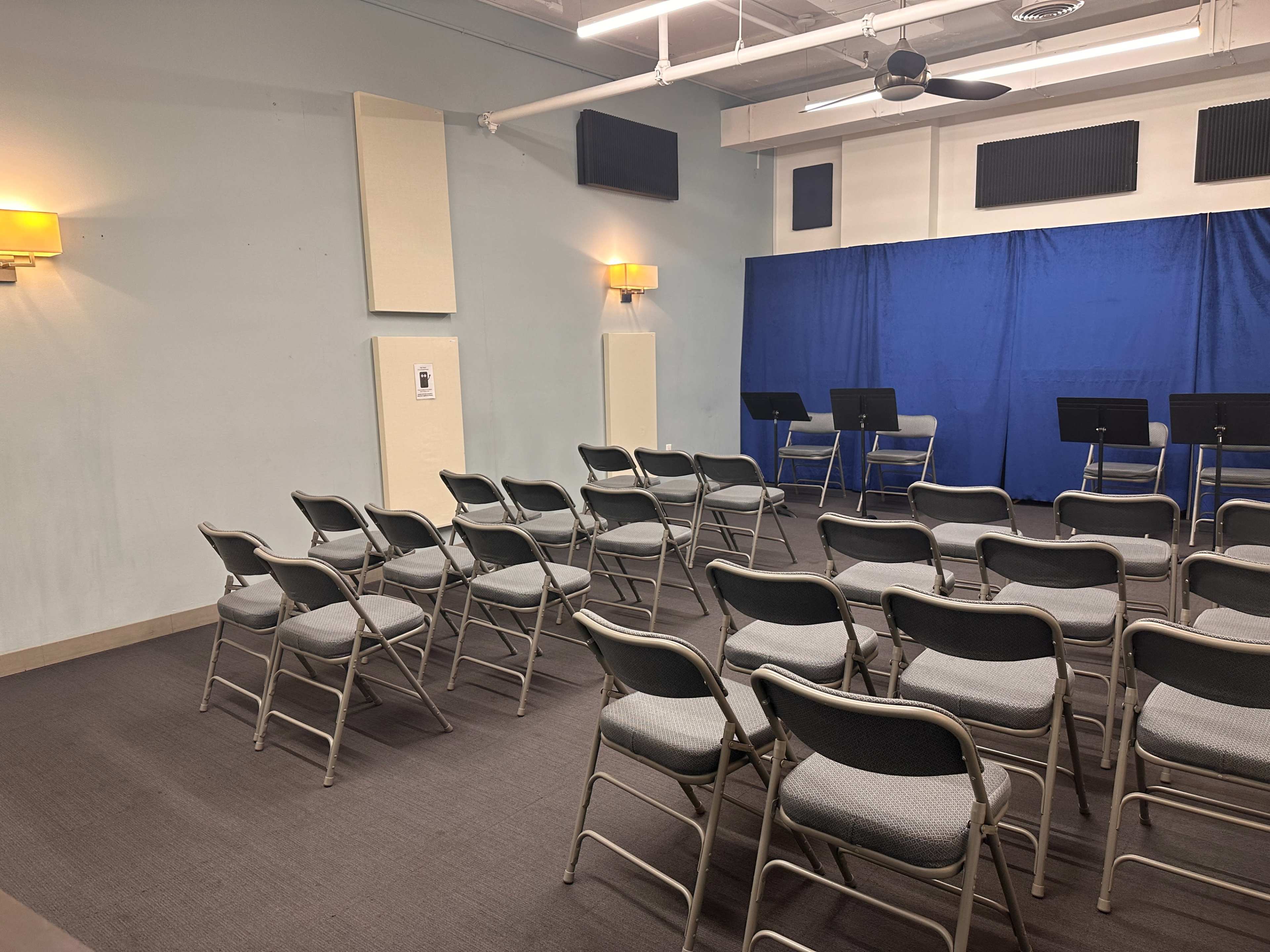 The image shows a small room arranged with folding chairs facing a blue backdrop and music stands.