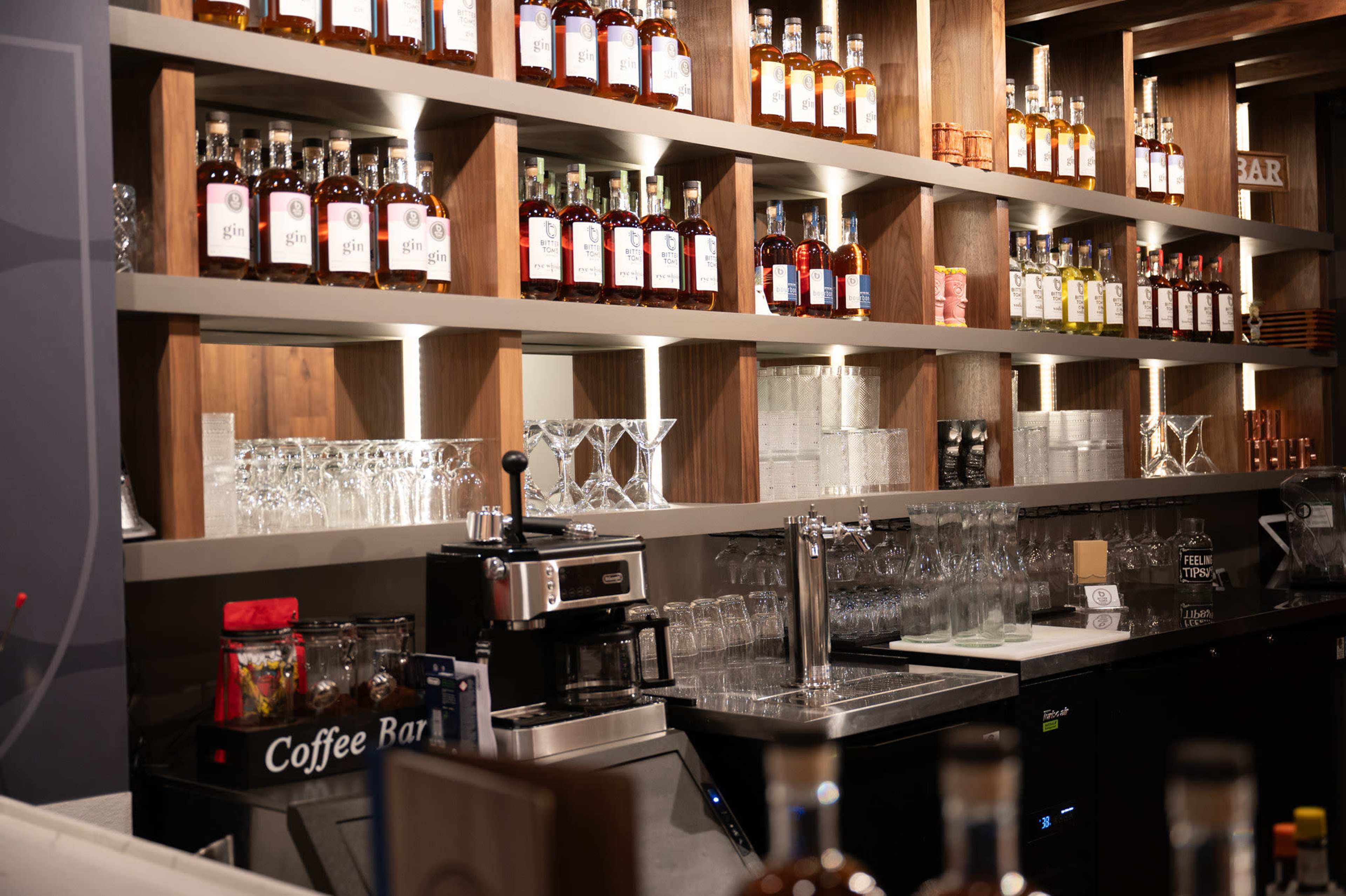 The image shows a well-organized bar with shelves lined with various bottles of liquor and a coffee bar setup in front.