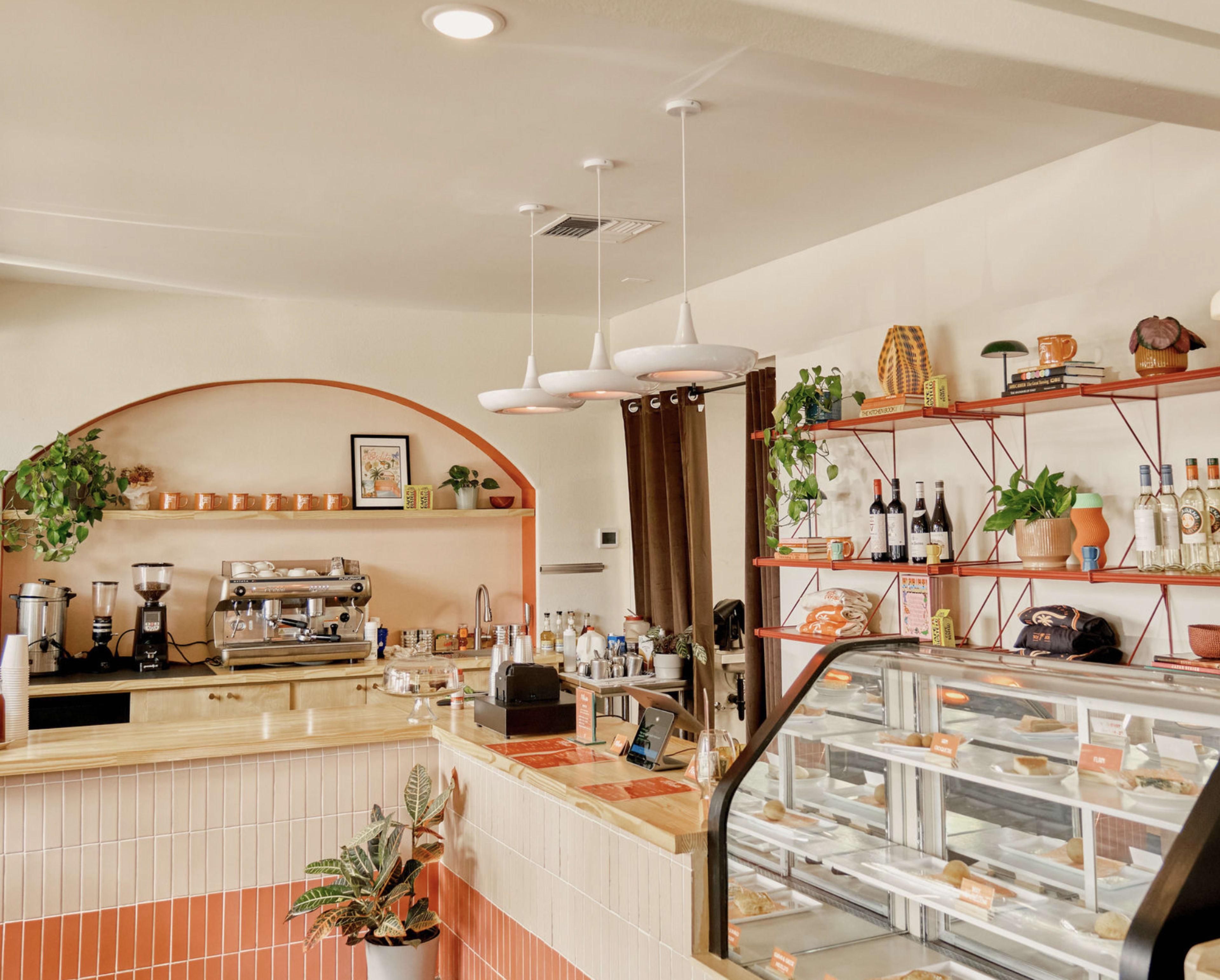 The image shows a modern café interior with a wooden counter, a coffee machine, and a display case filled with pastries, complemented by decorative plants and shelves showcasing bottles and pottery.
