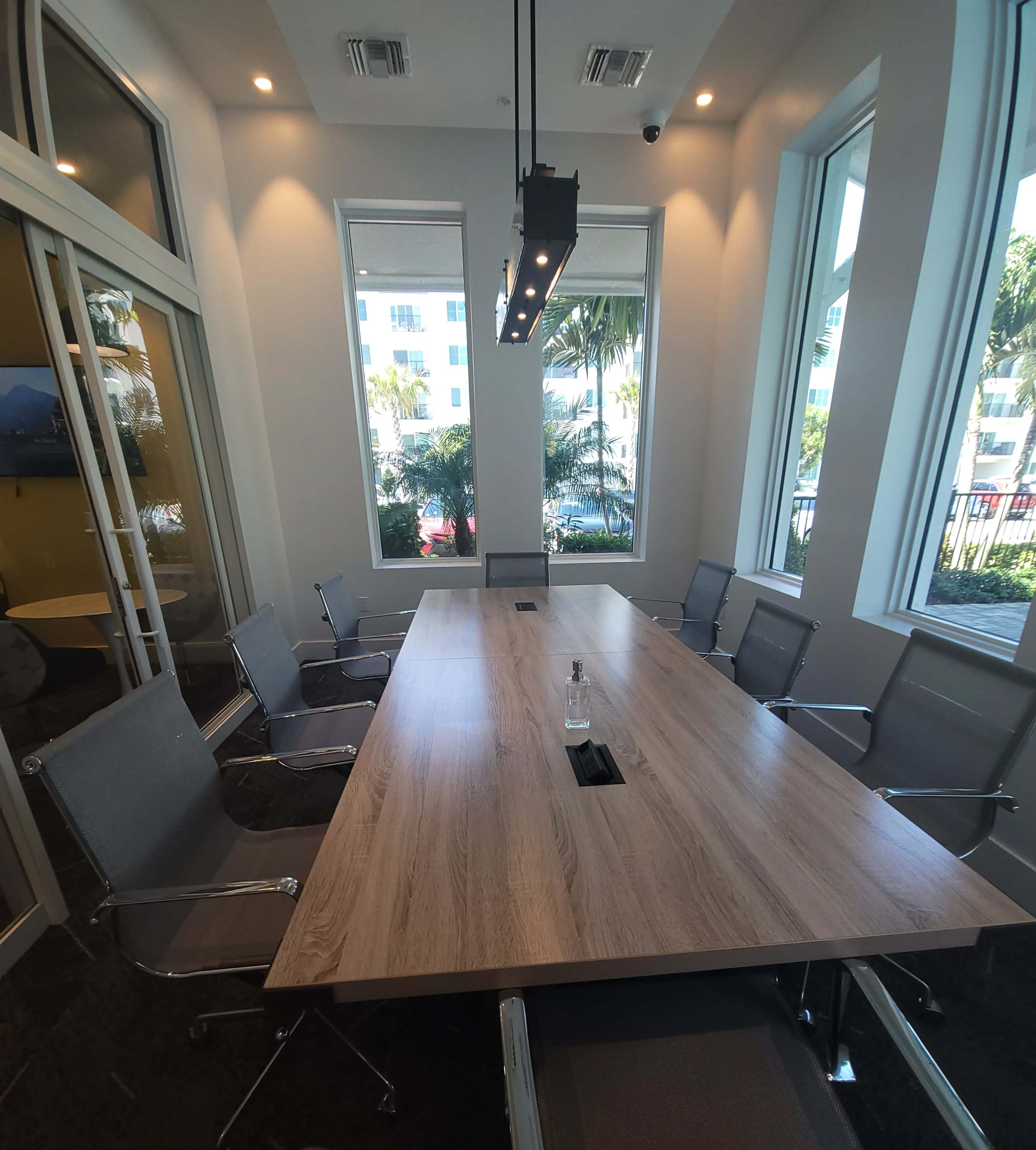 A modern conference room features a long wooden table surrounded by several metal-frame chairs, with large windows allowing natural light to enter.