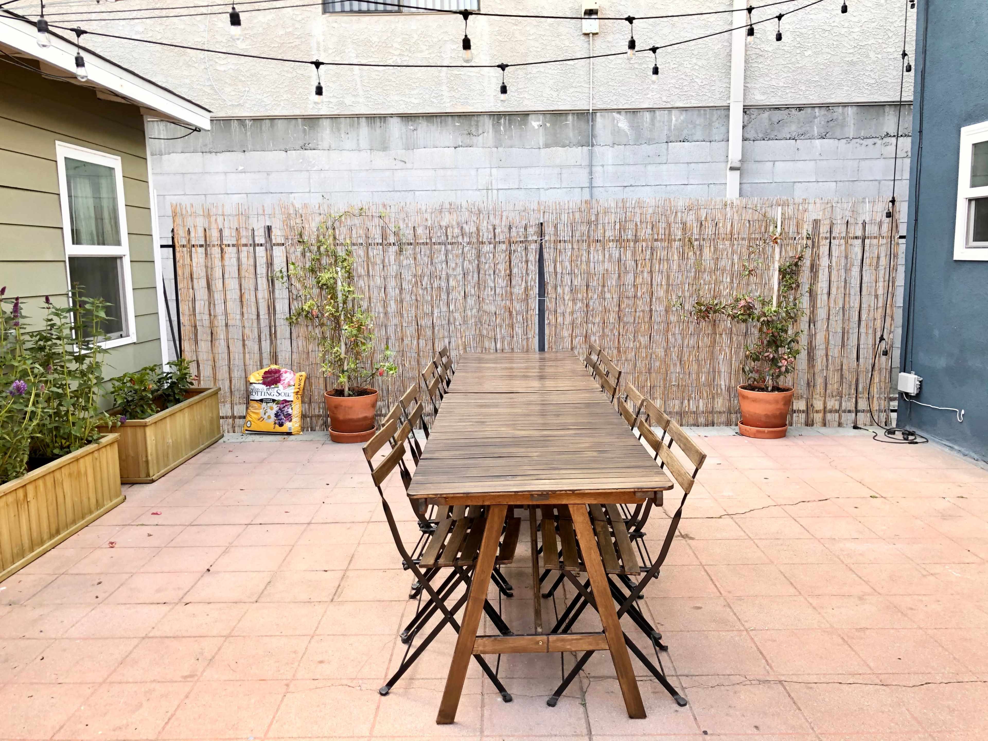 A long wooden table is set in a patio area, surrounded by folding chairs, with string lights overhead and planters flanking the space.