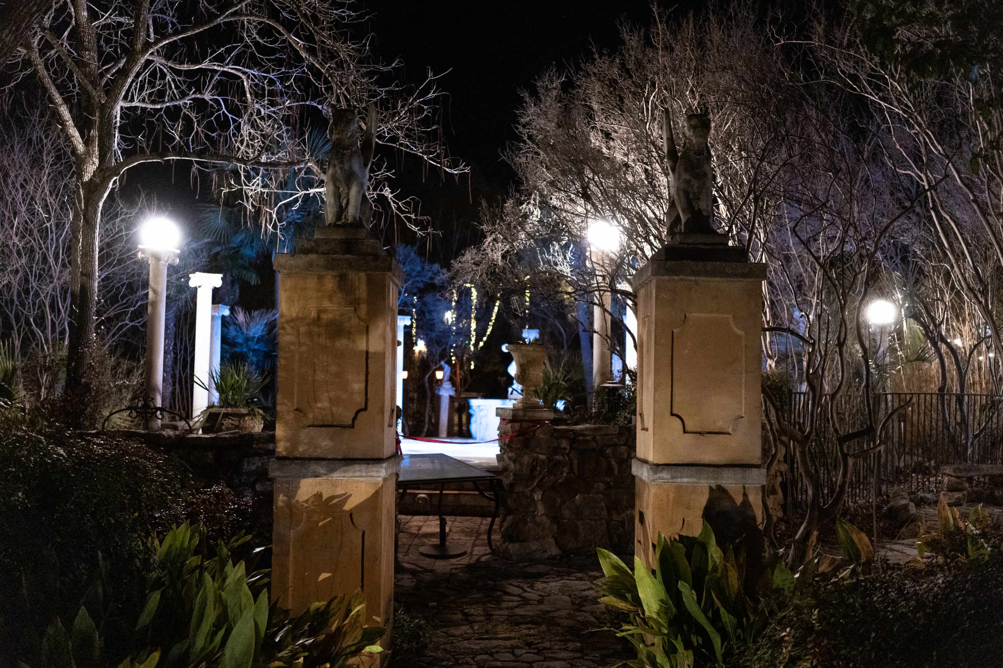 A stone entrance with lion sculptures leads to a dimly lit garden featuring a fountain and illuminated trees.