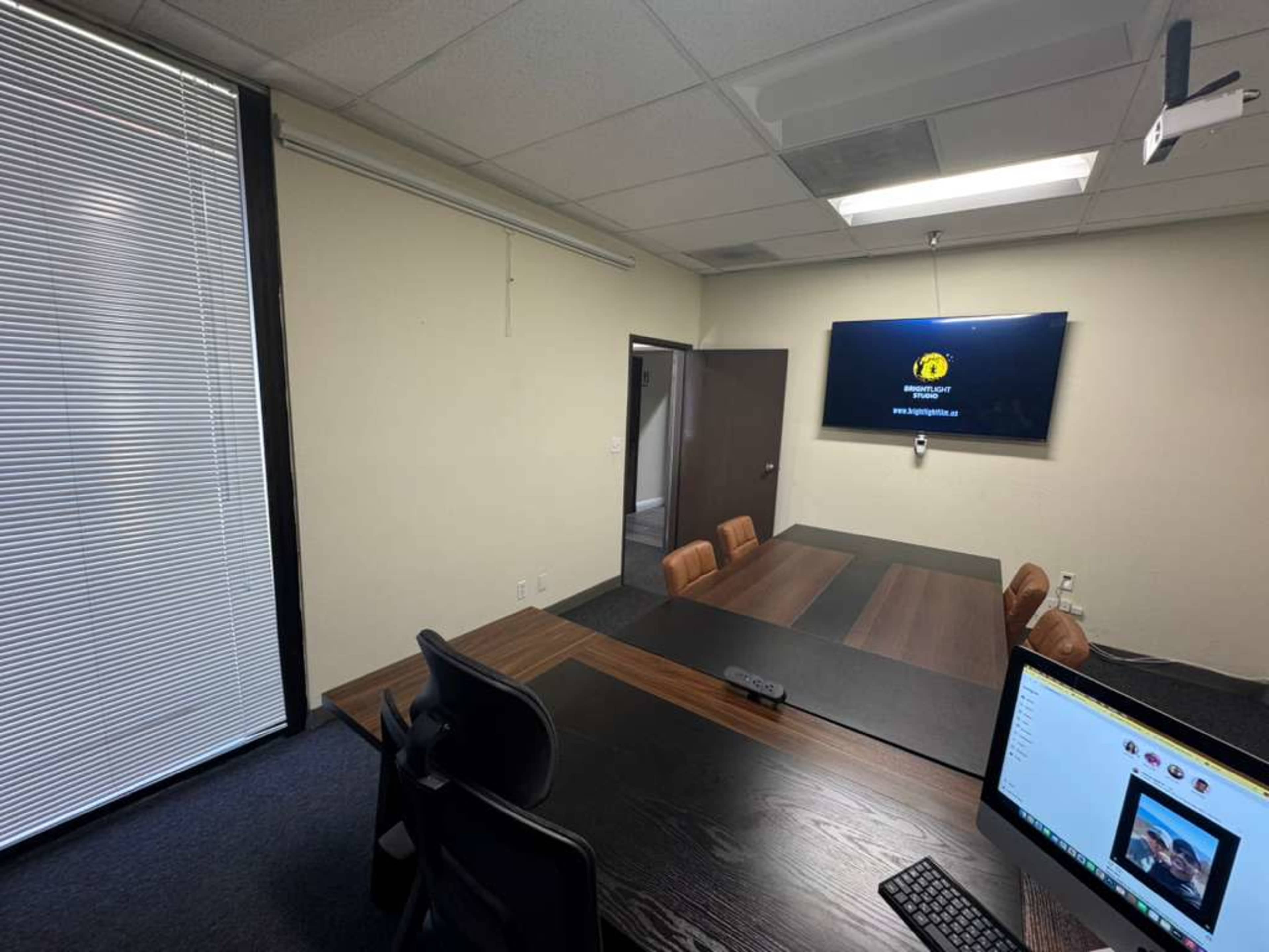 A conference room features a large wooden table with several chairs, a wall-mounted screen, and a window with blinds.