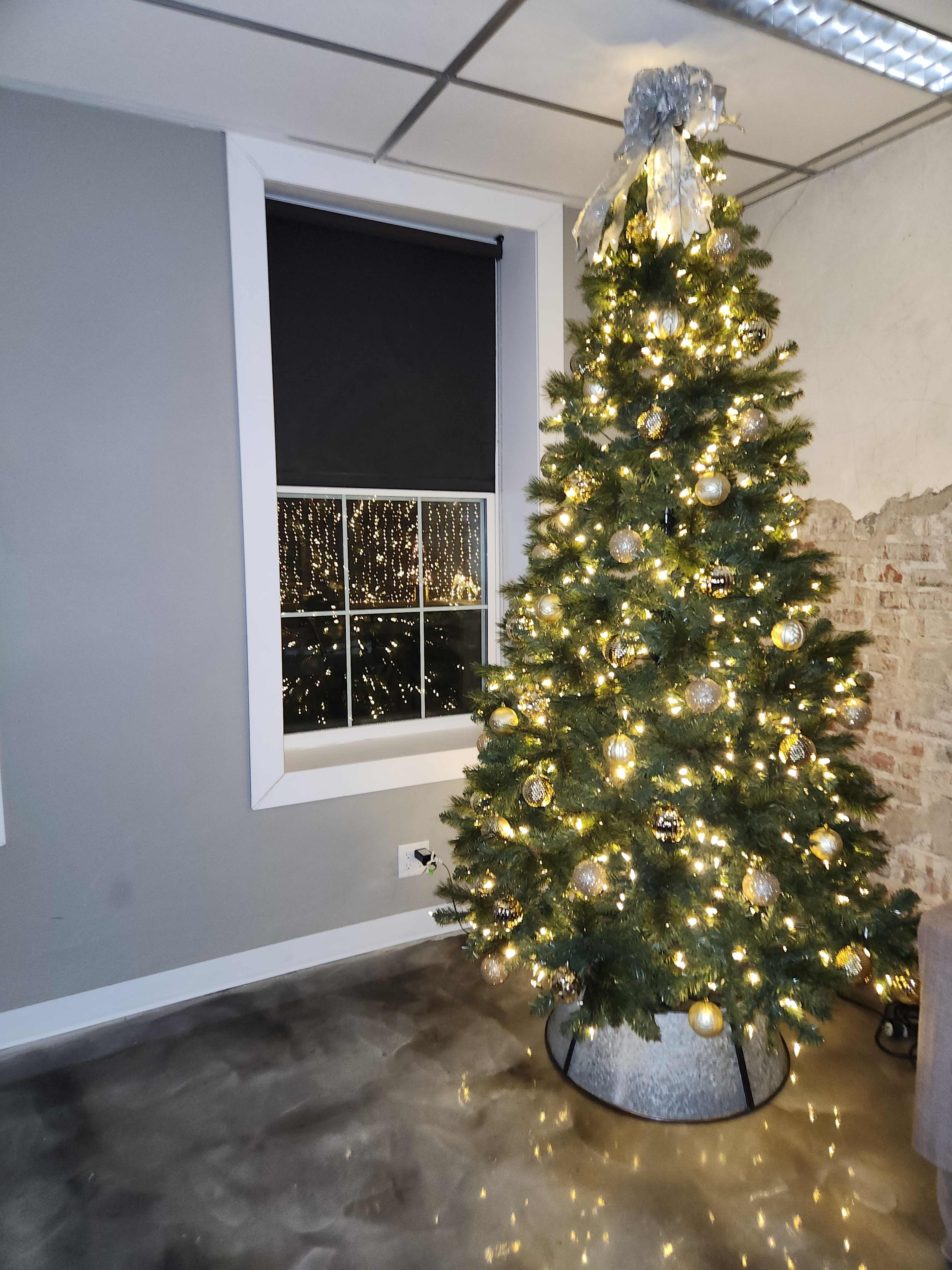 A decorated Christmas tree with white lights and ornaments stands in a room next to a window with a view of city lights reflected in the glass.