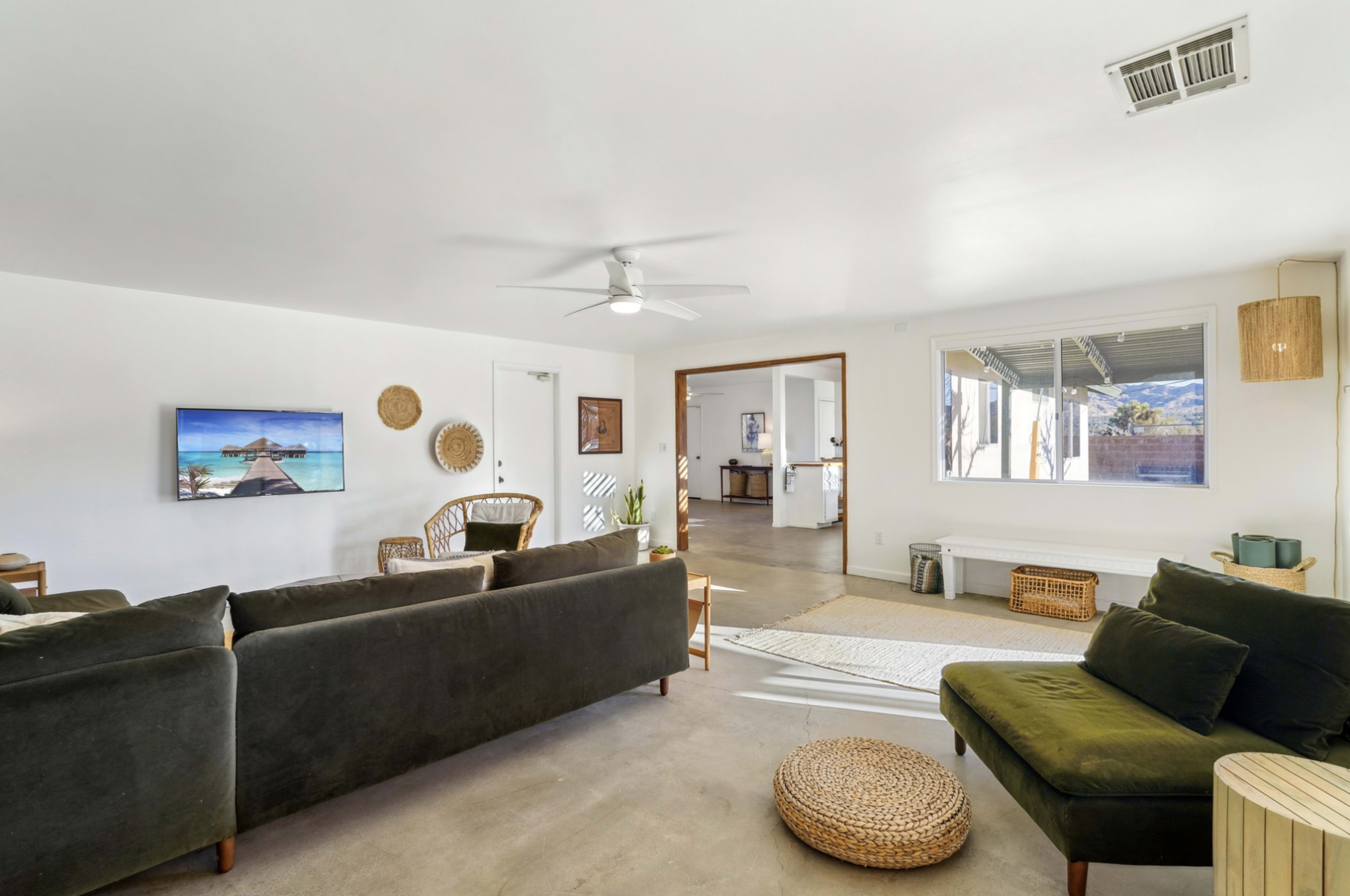 The image features a modern living room with a dark green sofa, a television mounted on the wall, and a view into an adjacent room with a kitchen area.