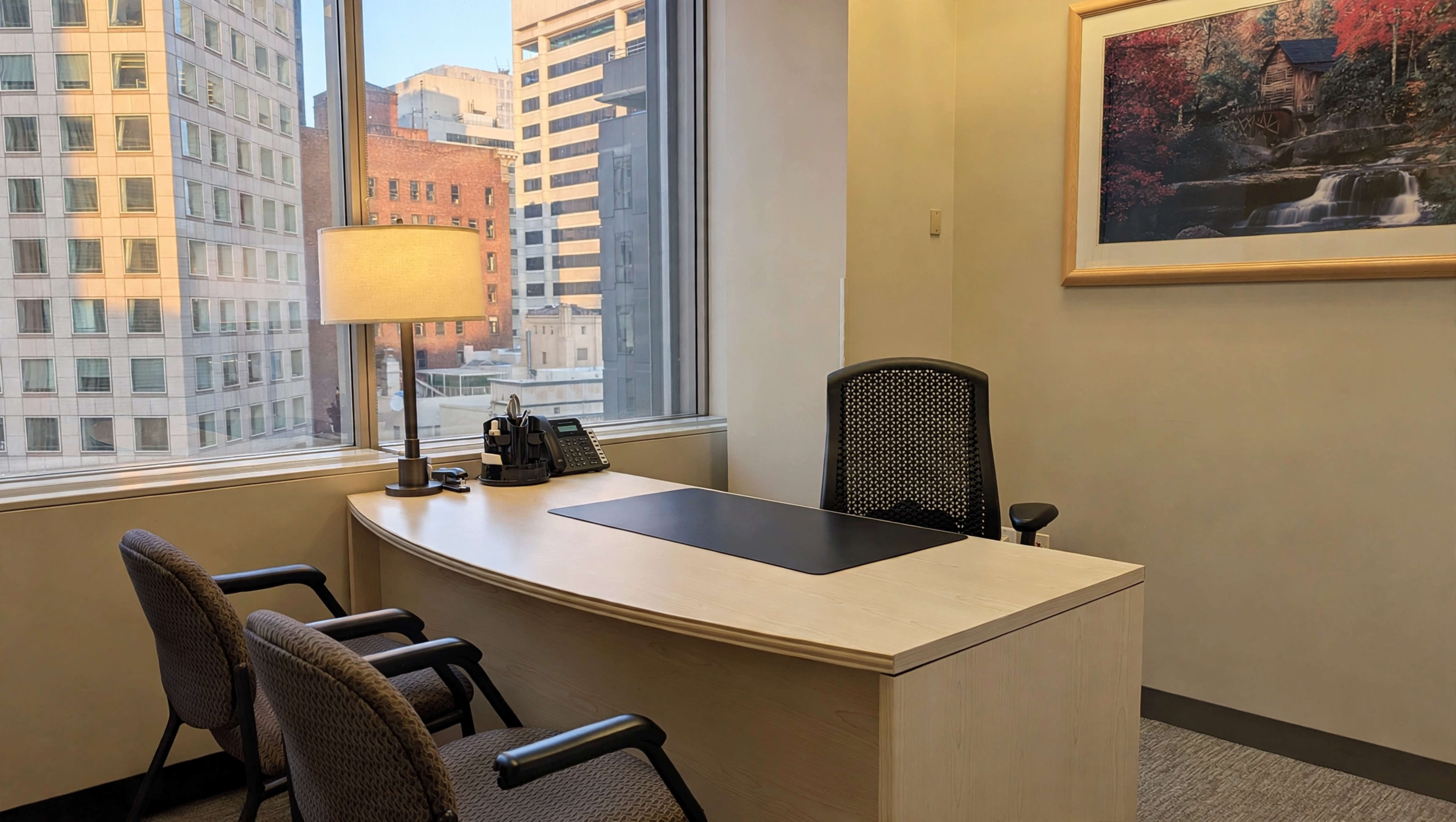 An office space features a desk, a lamp, and two chairs, with large windows showcasing a view of city buildings.