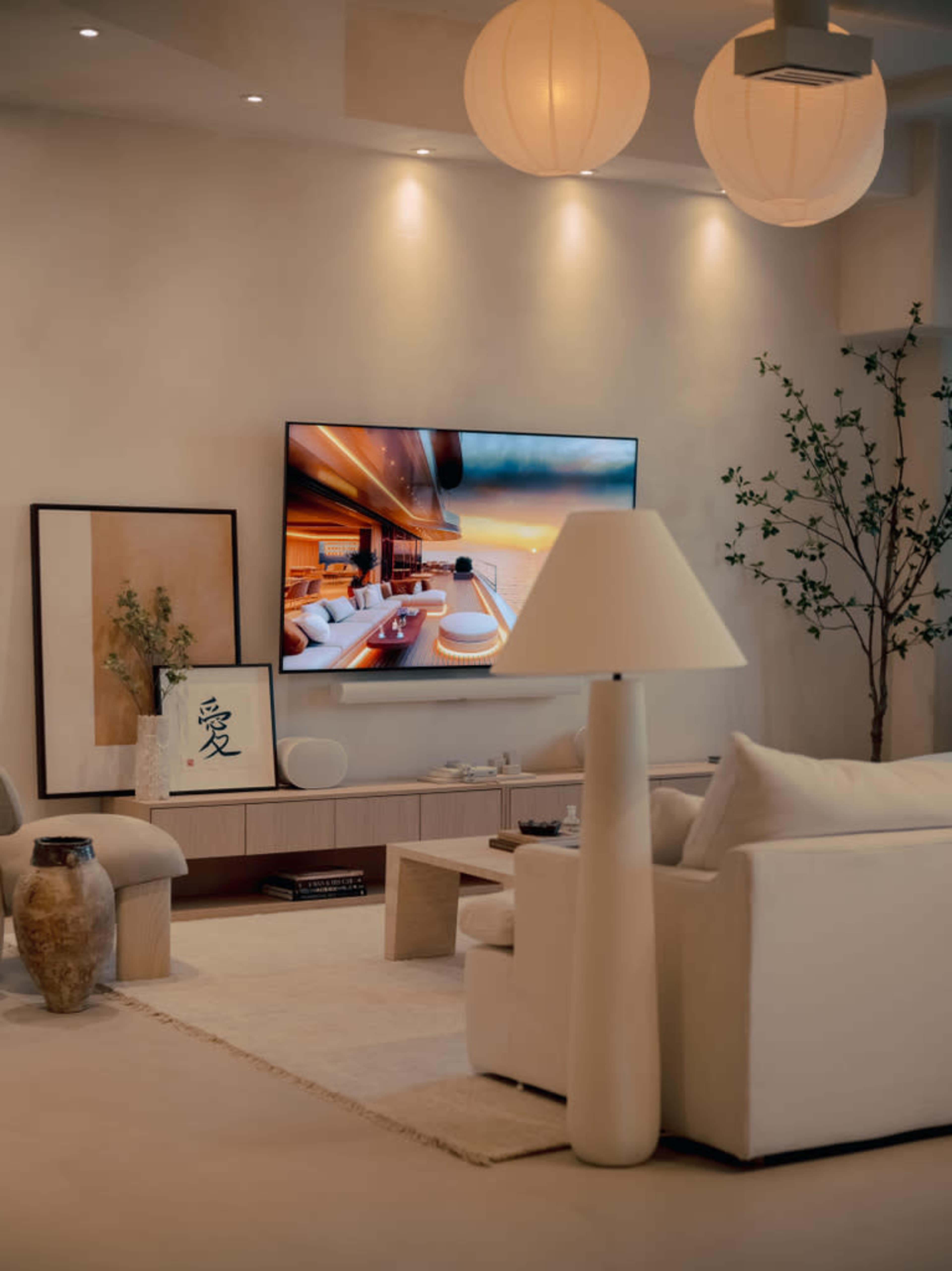 A modern living room features a light-colored sofa, a large TV mounted on the wall displaying a scenic image, and decorative items including a lamp and artwork.