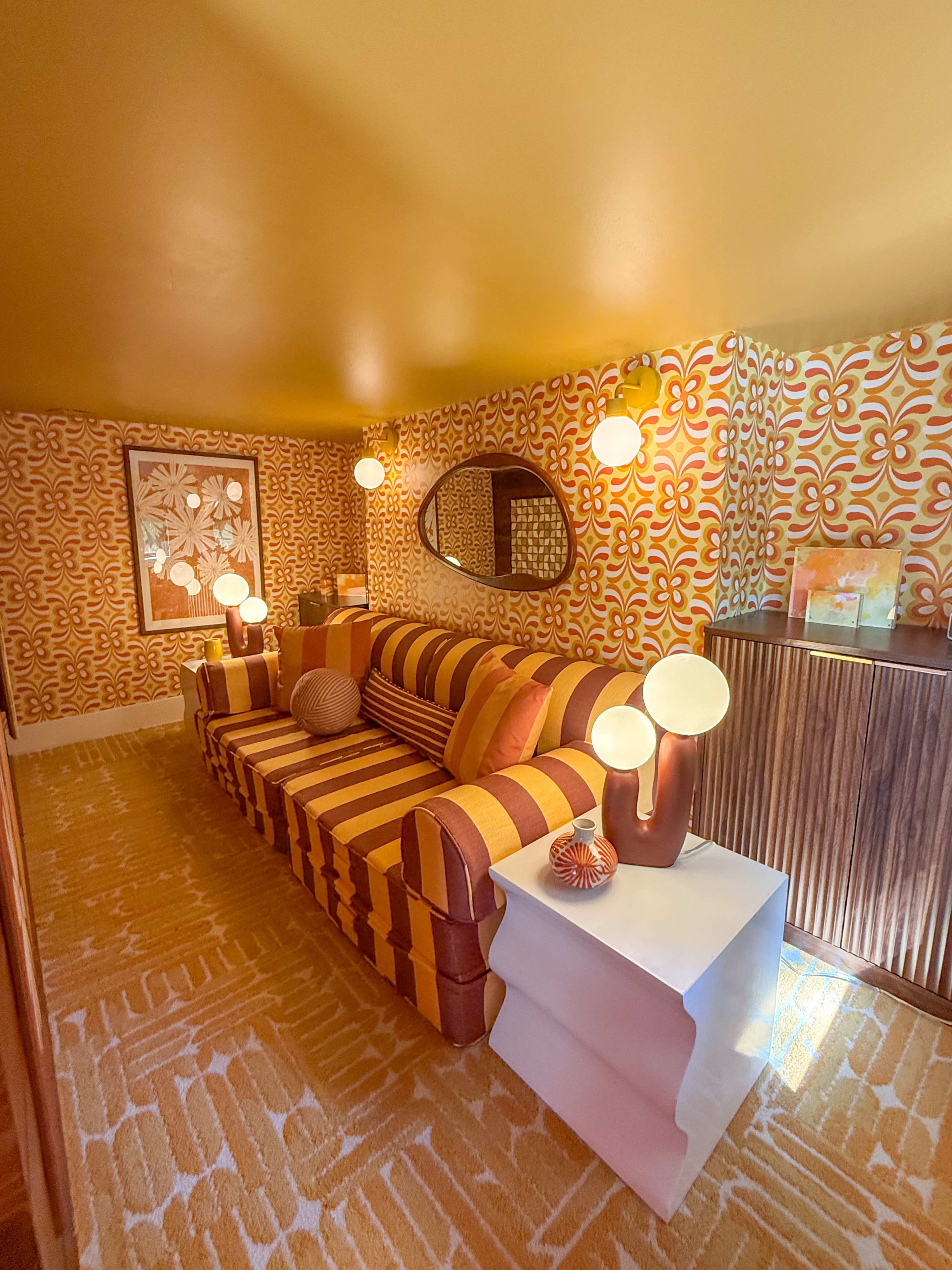 The image shows a retro-styled living room with orange and yellow patterned wallpaper, a striped sofa, and round pendant lights.