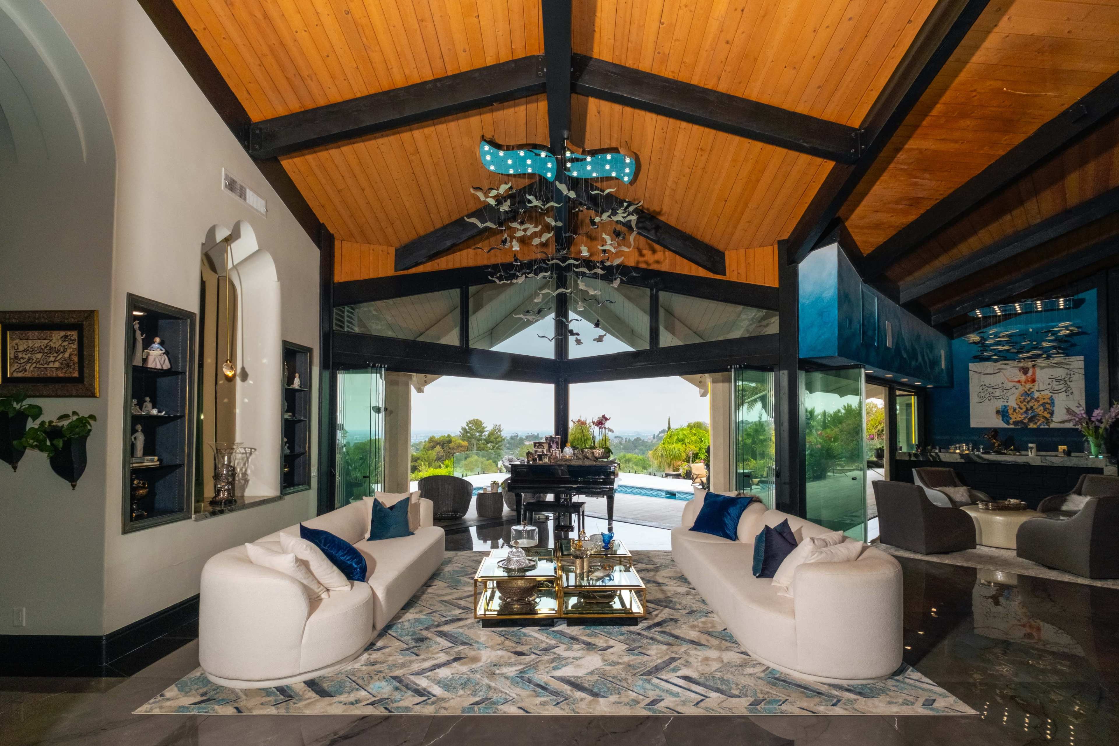 The image shows a modern living room with high wooden ceilings, large glass windows overlooking a pool, and contemporary furniture arranged around a central coffee table.