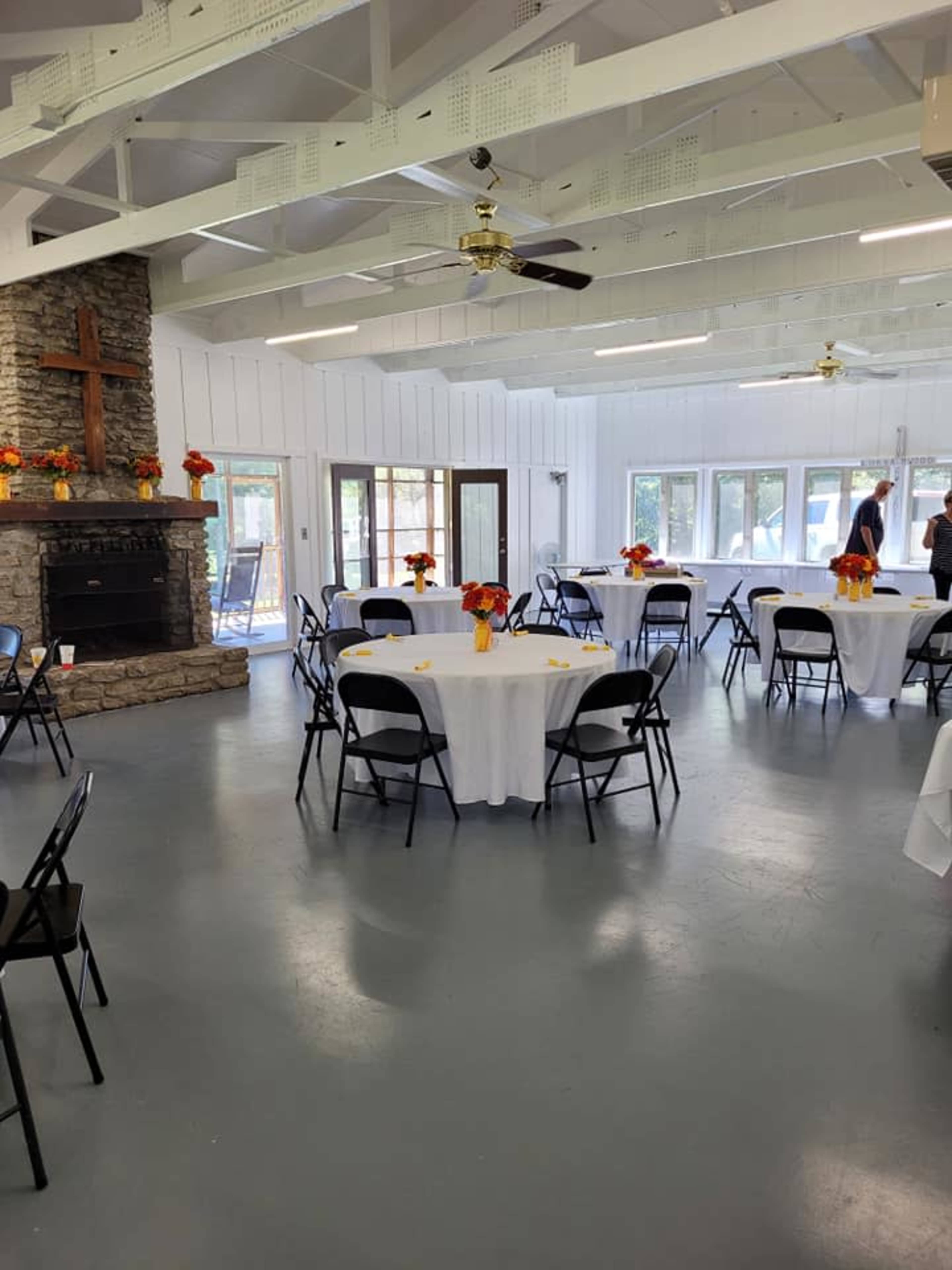 A spacious indoor venue with white walls, a stone fireplace featuring a cross, round tables covered with white tablecloths, and chairs arranged around them, all decorated with floral centerpieces.