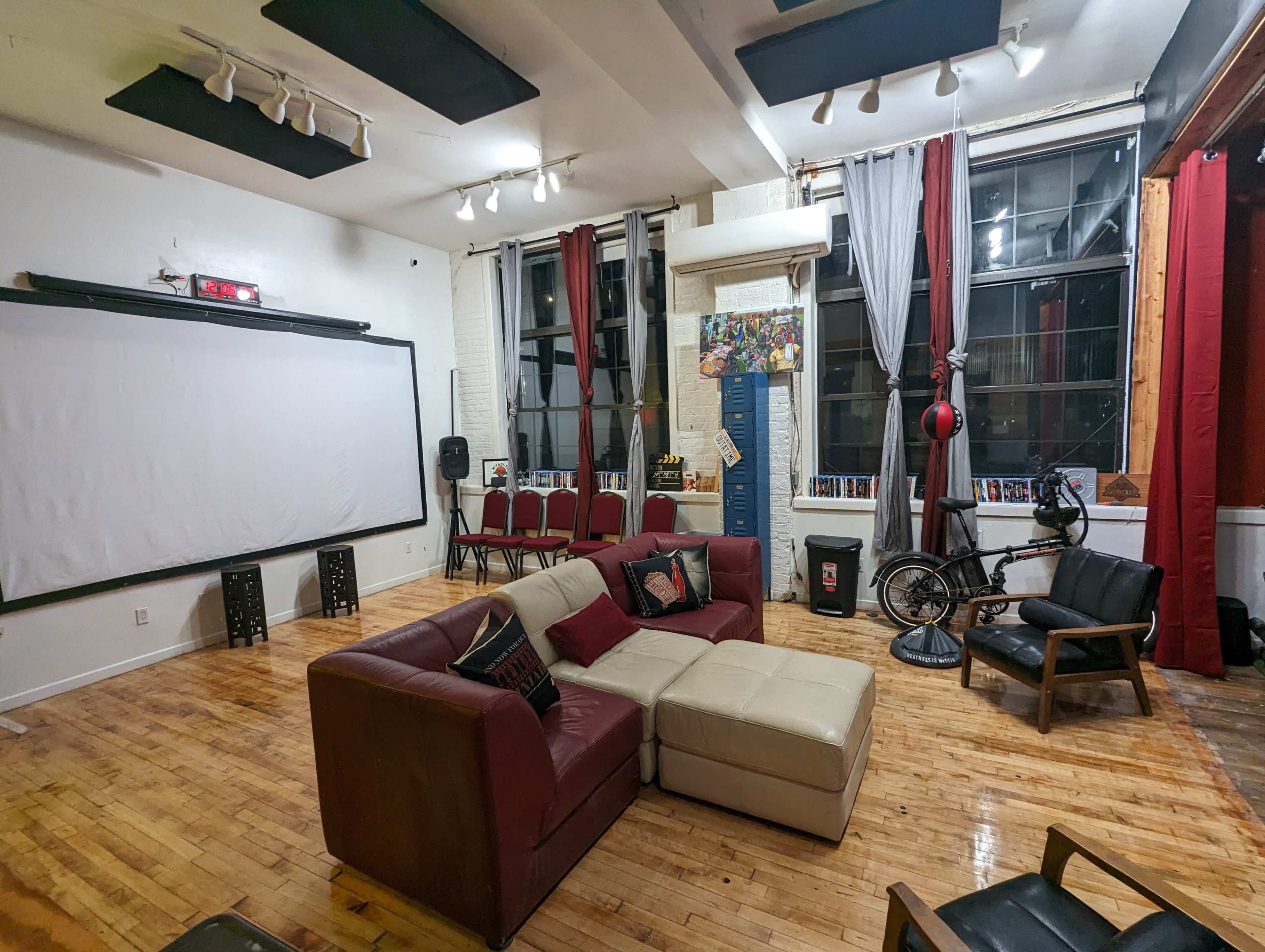 A spacious room with a large screen on one wall, seating arranged around a central couch, and various decorative elements including a bicycle and colorful curtains.