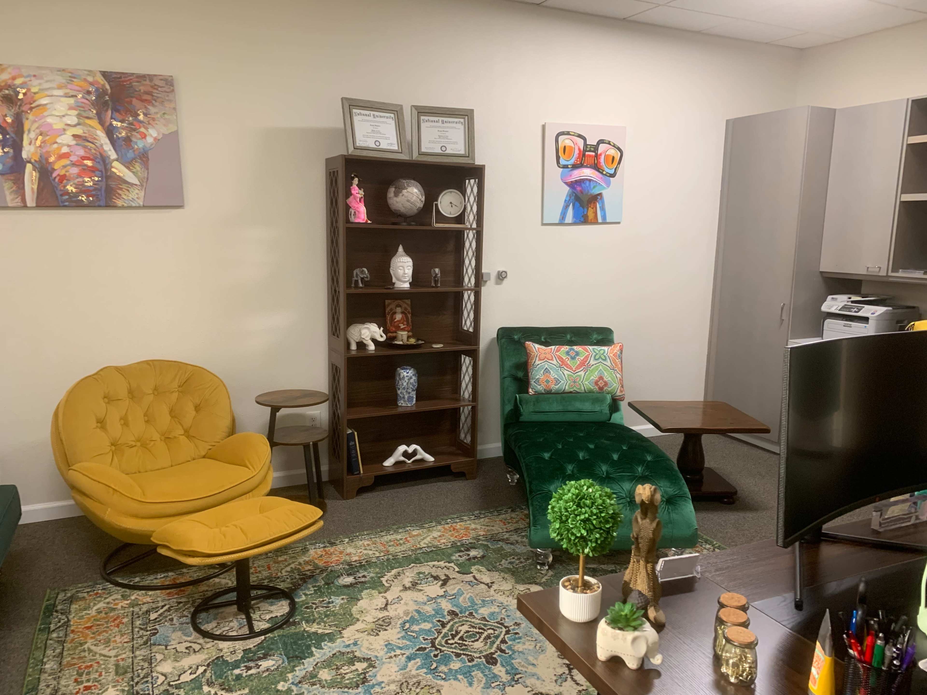 The image shows a room furnished with a yellow chair, a green chaise lounge, a bookshelf with decorative items, and a desk with a computer and various office supplies.