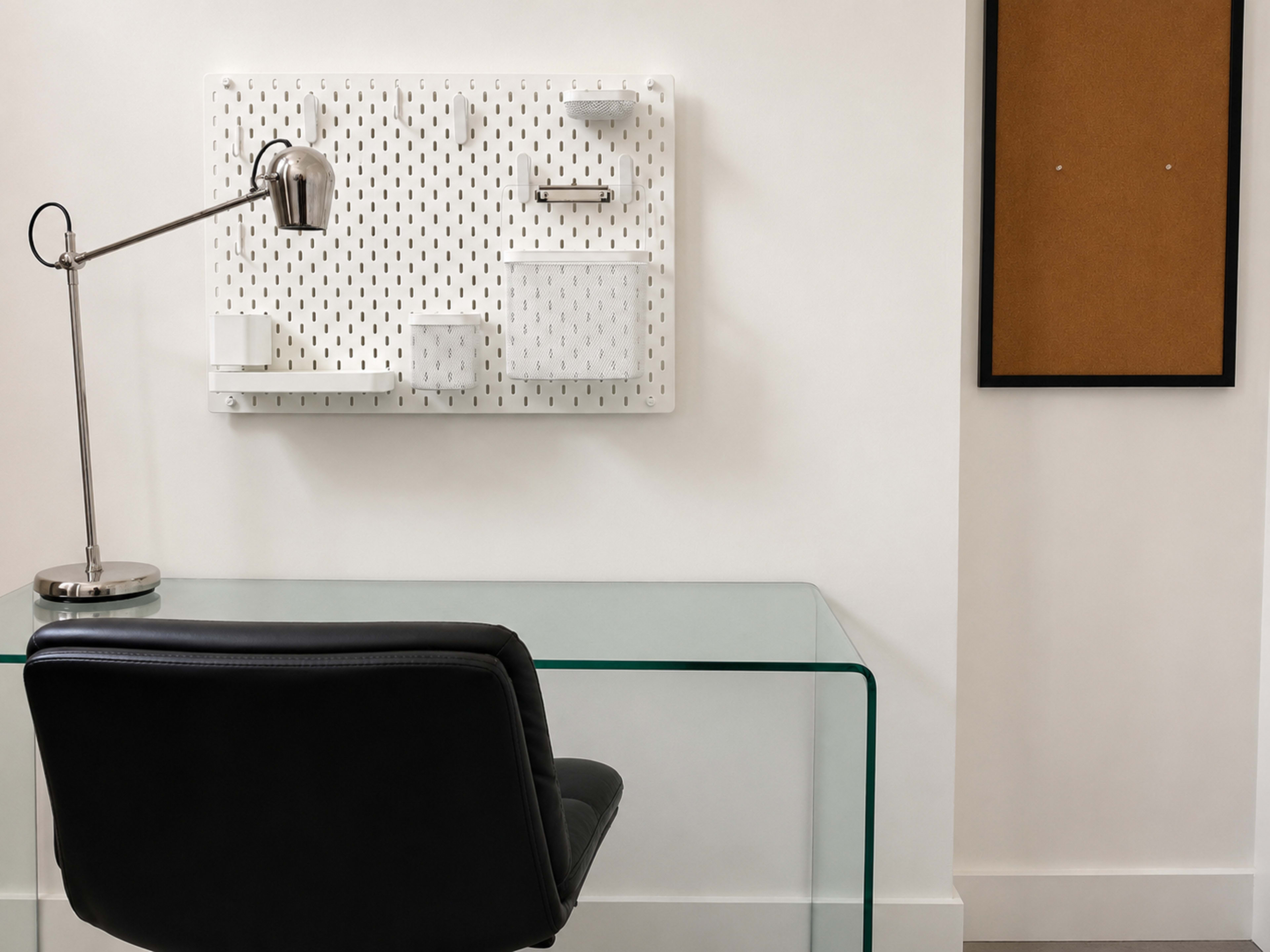 A modern workspace features a clear glass desk with a black chair, accompanied by a wall-mounted organizer and a corkboard in a minimalist setting.