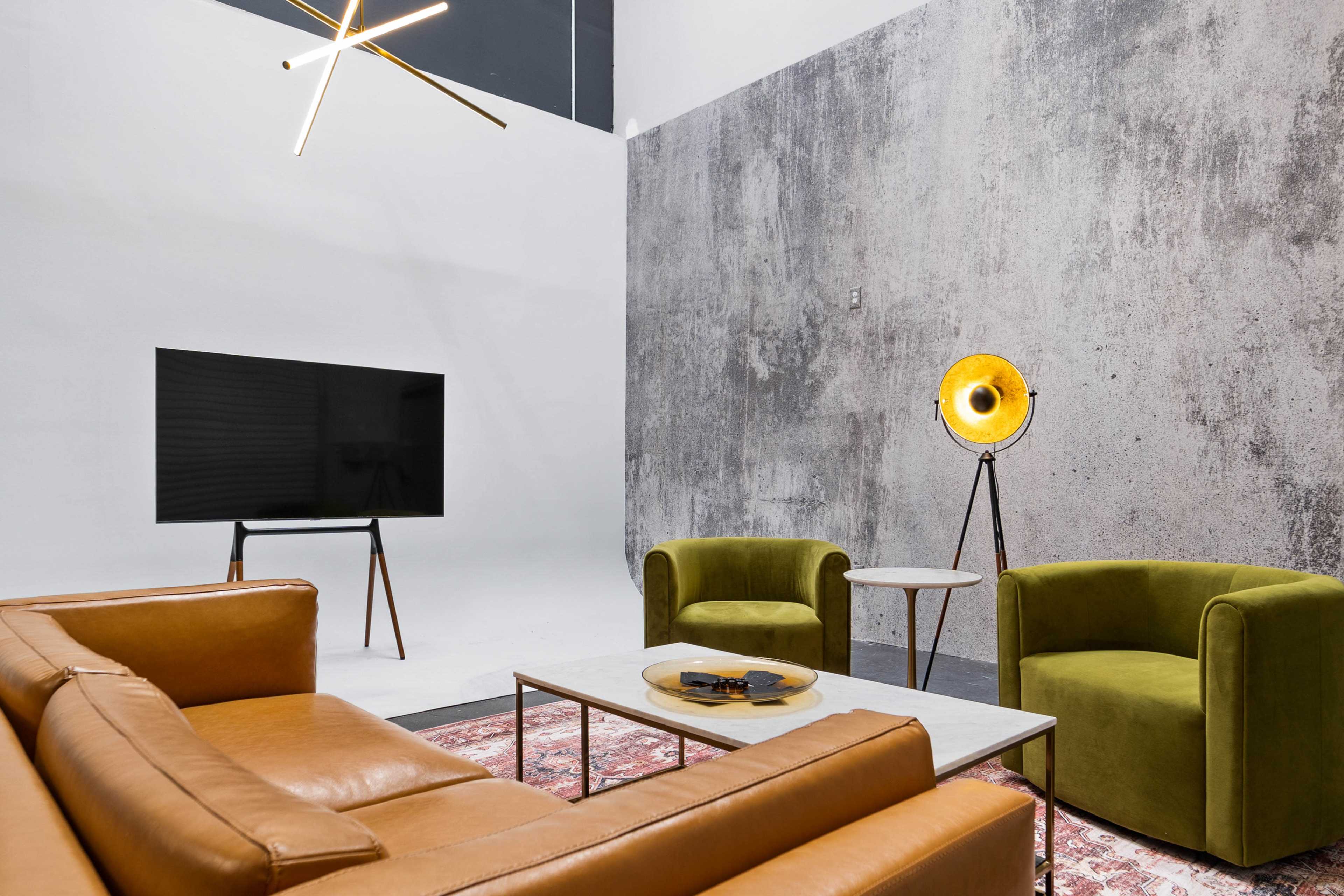 The image features a modern living space with a brown leather sofa, two green armchairs, a marble coffee table, a floor lamp, and a television against a textured gray wall.