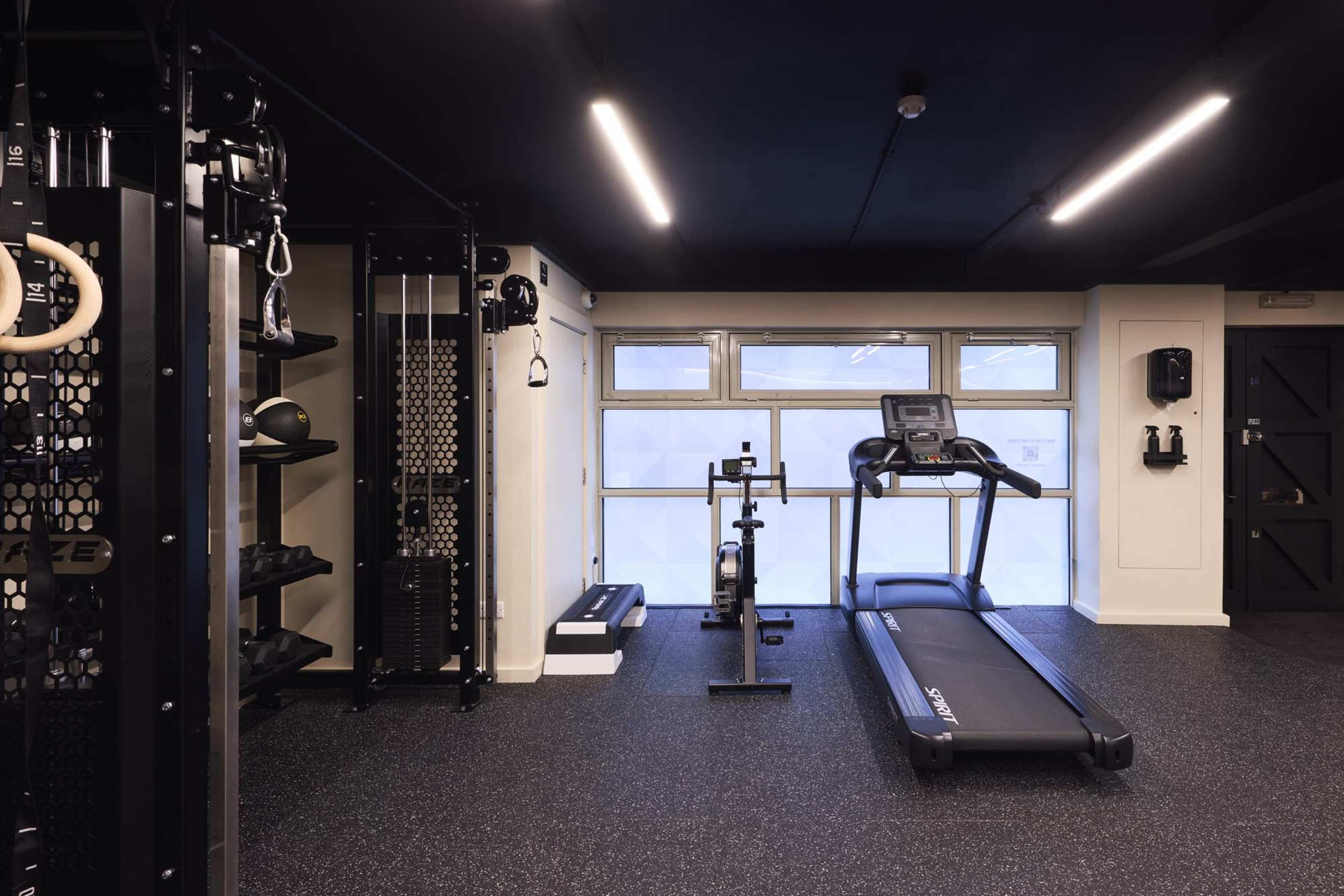 The image shows a modern gym space featuring a treadmill, a stationary bike, and various exercise equipment against a dark wall and floor.