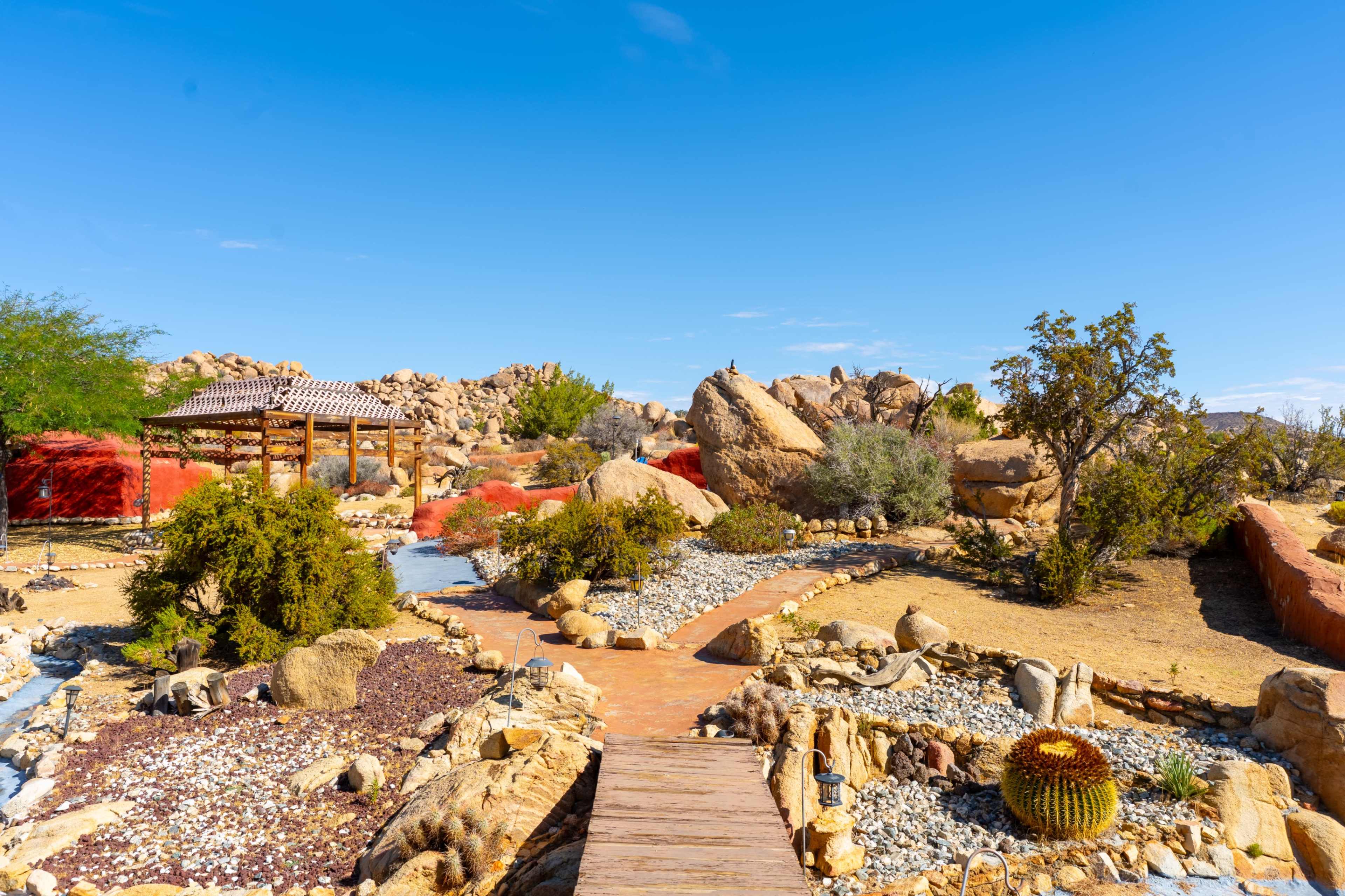 The scene features a desert landscape with rock formations, a wooden walkway, and a gazebo surrounded by various plants and stones.