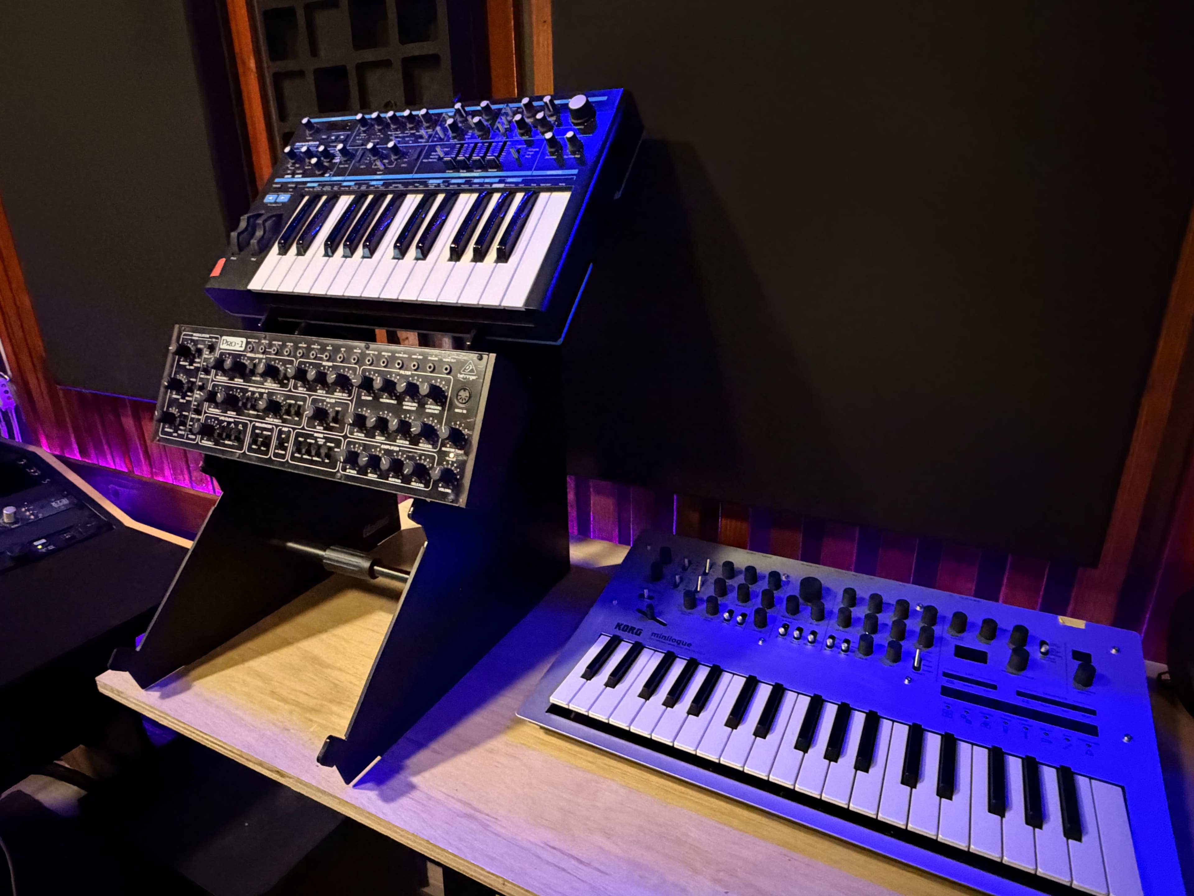The image displays three synthesizers arranged on a wooden stand in a music studio, with purple LED lighting in the background.