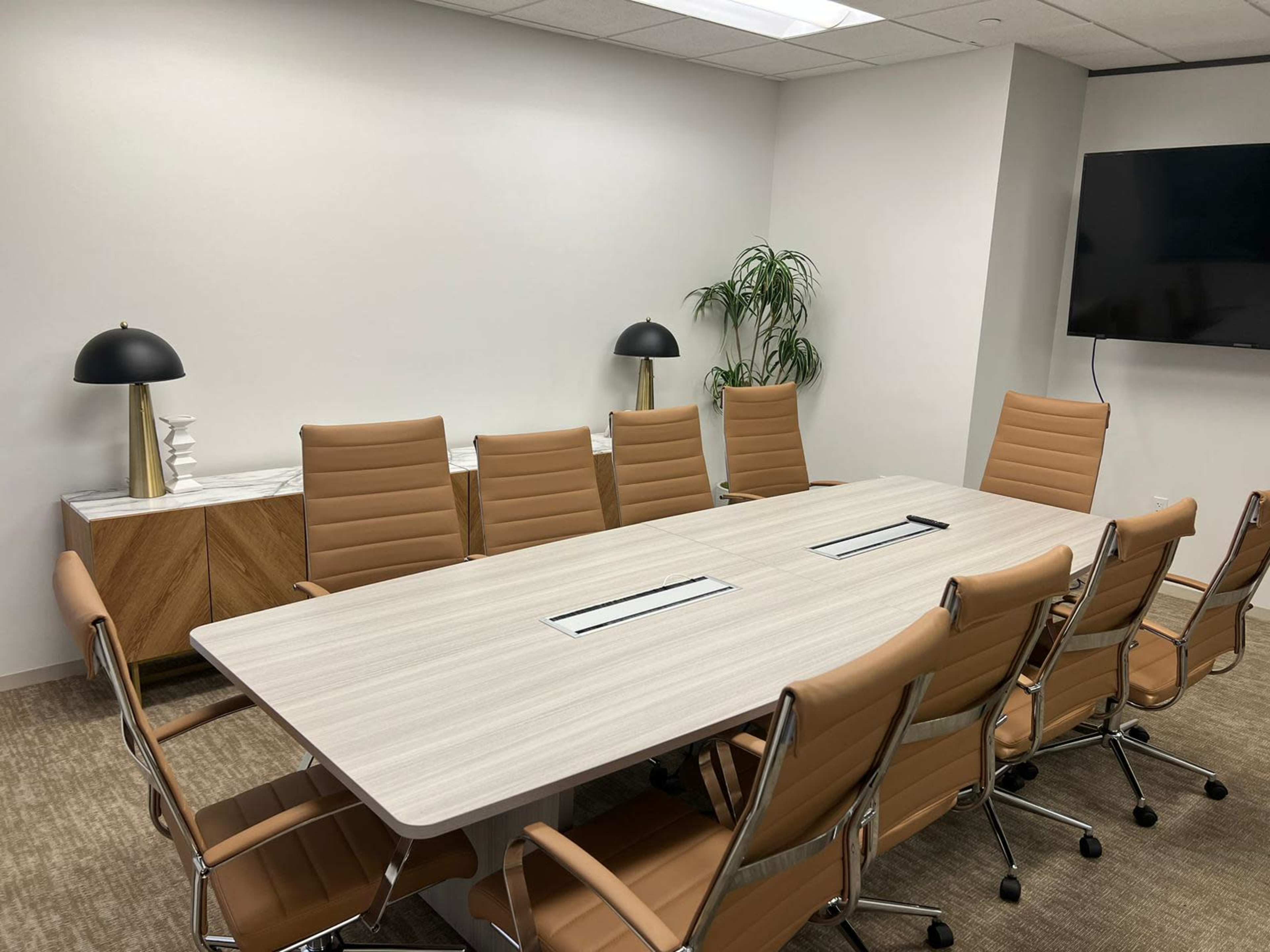 A modern conference room with a long table surrounded by eight brown chairs, against a backdrop of a sideboard and a TV on the wall.