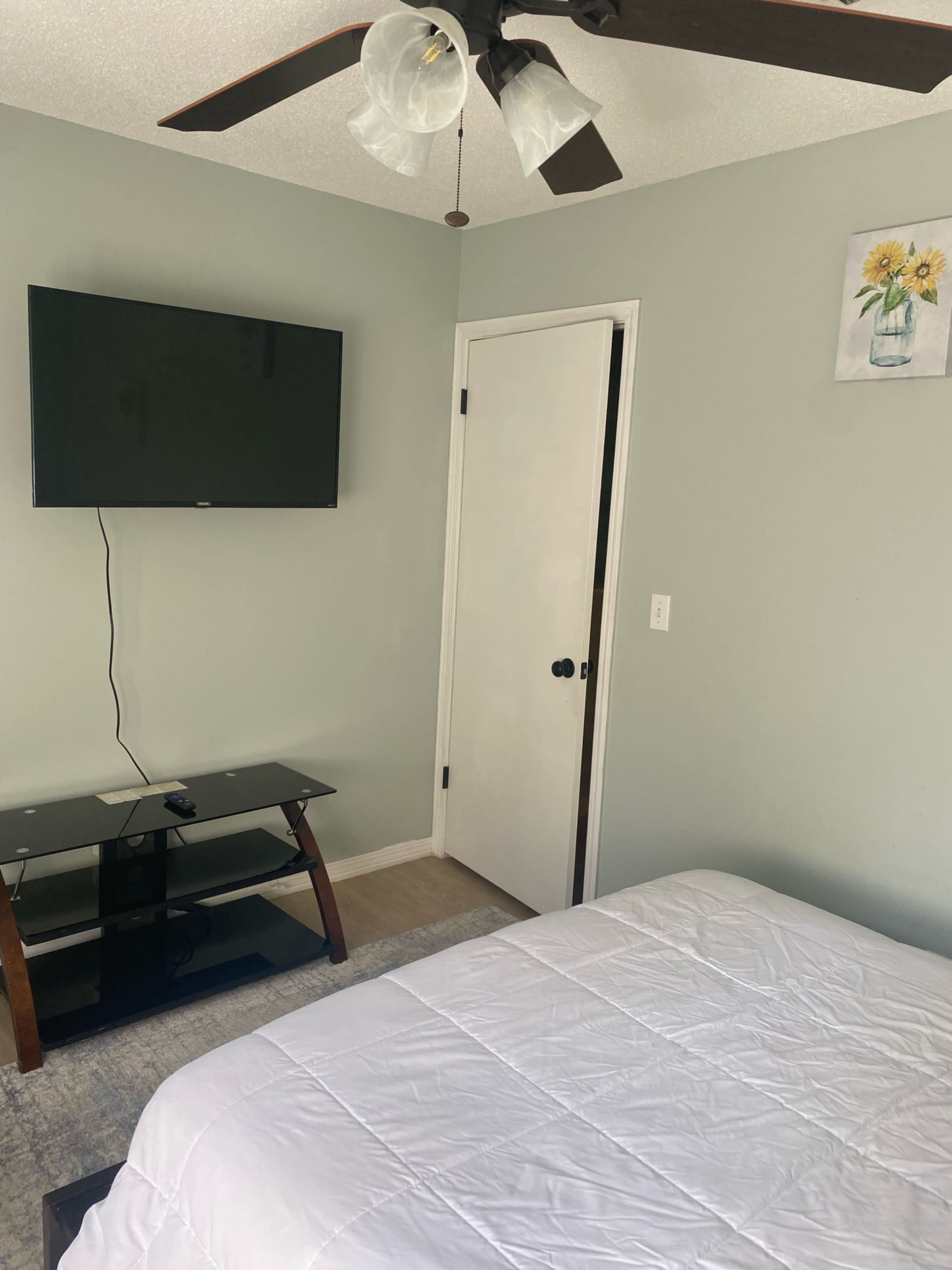 A bedroom with a television mounted on the wall, a glass TV stand, a white bedspread, and a closed door.