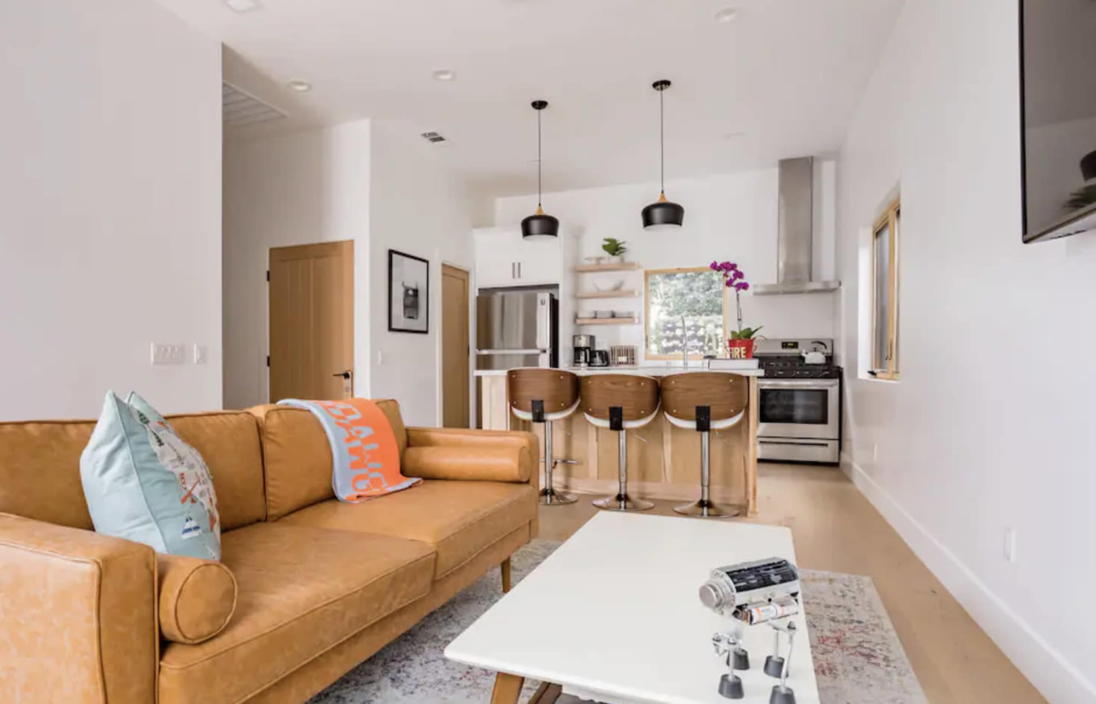 A modern living space features a tan leather sofa, a light coffee table, and a kitchen area with bar stools and stainless-steel appliances.