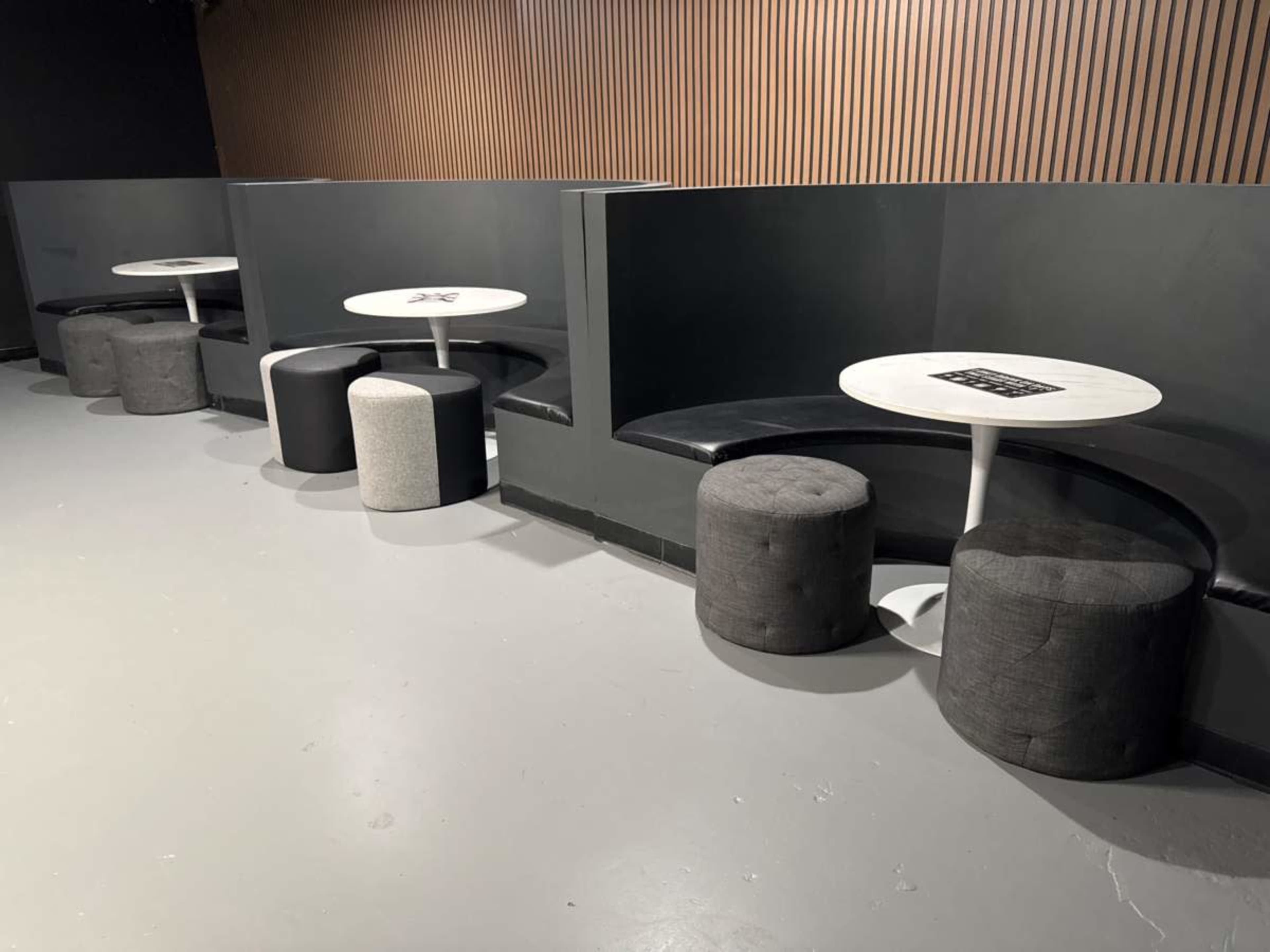 The image shows a modern seating area with several small round tables and cushioned stools set against wooden-paneled walls.