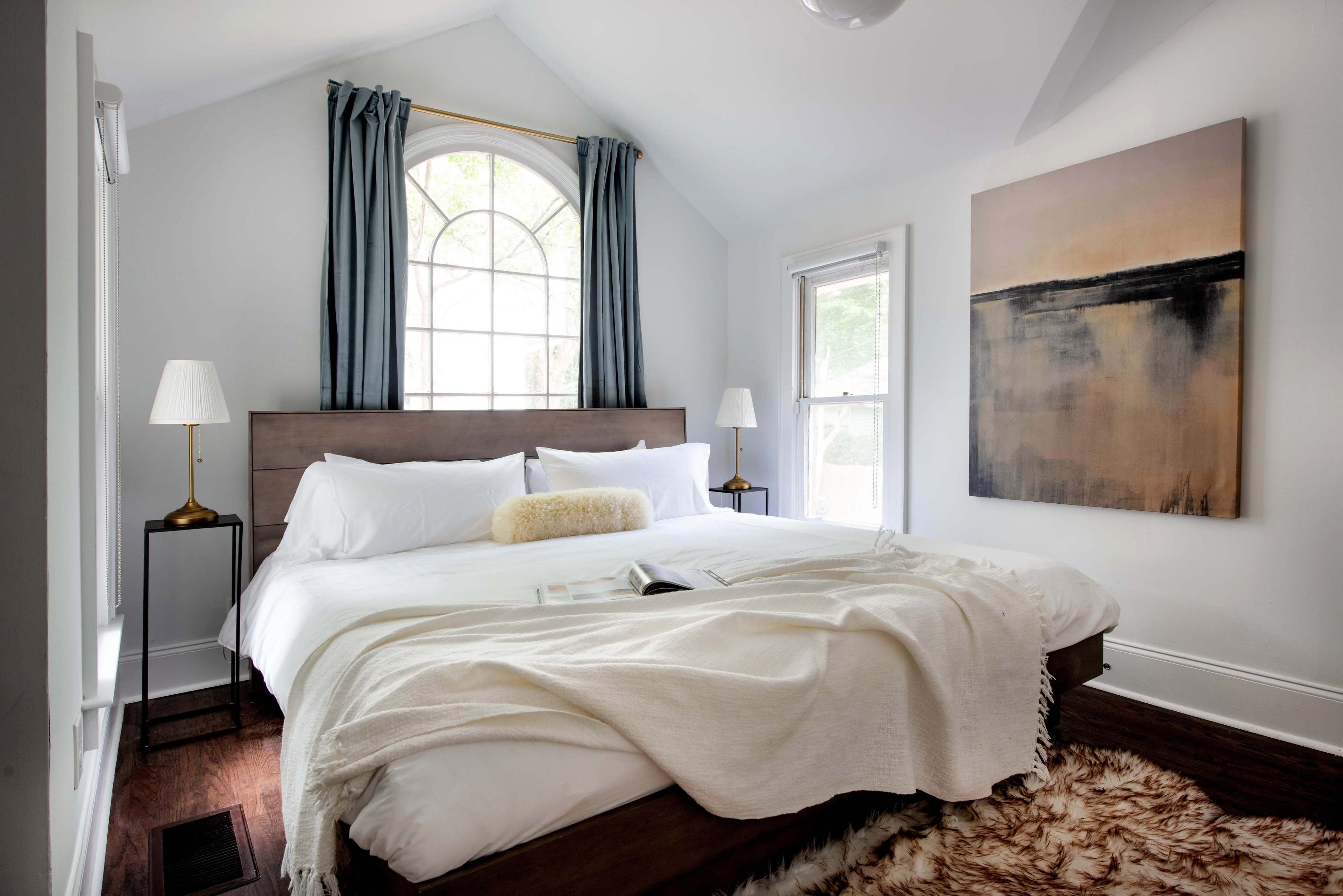 A neatly arranged bedroom features a large bed with white bedding, two bedside lamps, and a large window with sheer curtains.
