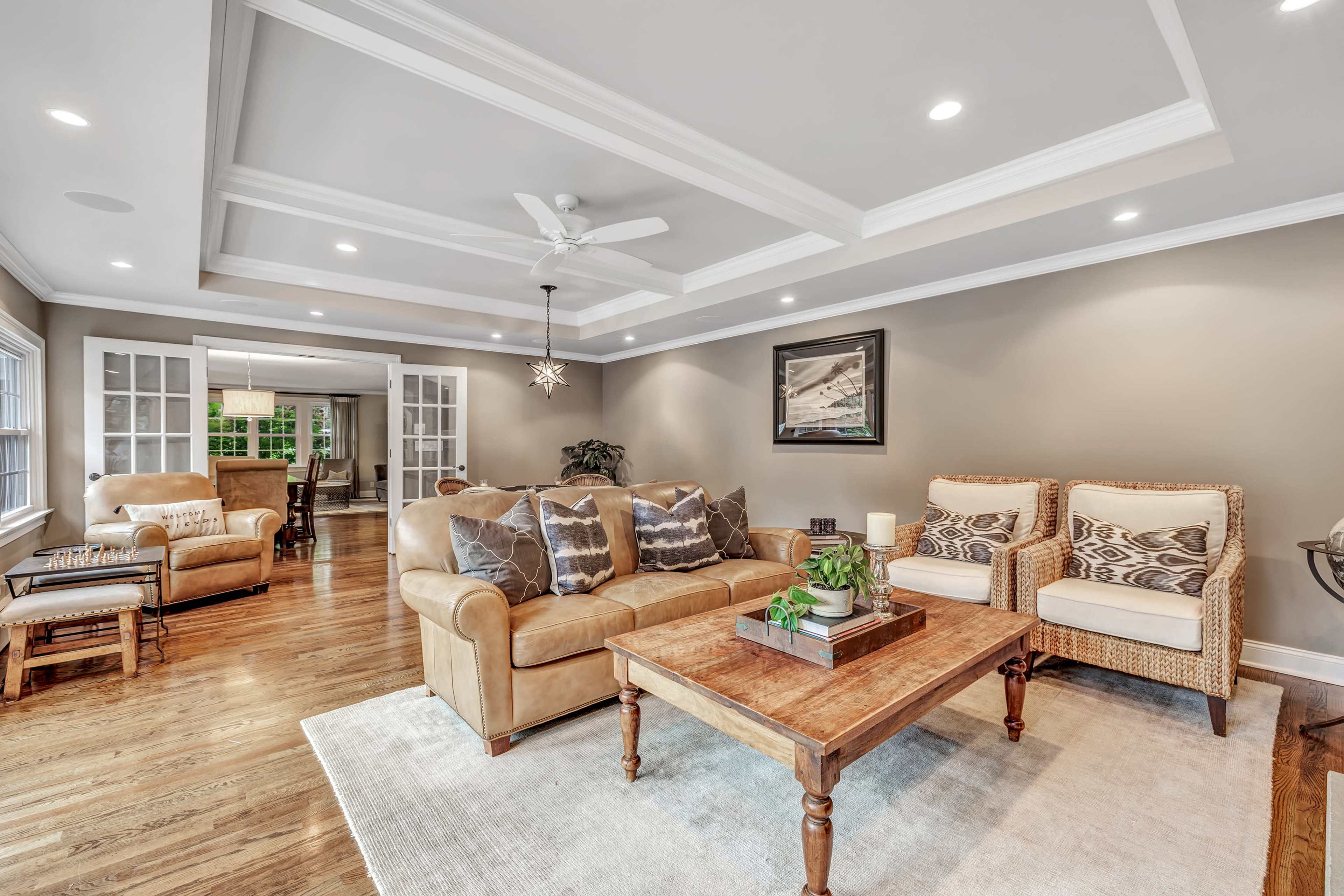 A well-lit living room features a beige couch, two patterned armchairs, a wooden coffee table, and large windows with a view into an adjoining dining area.