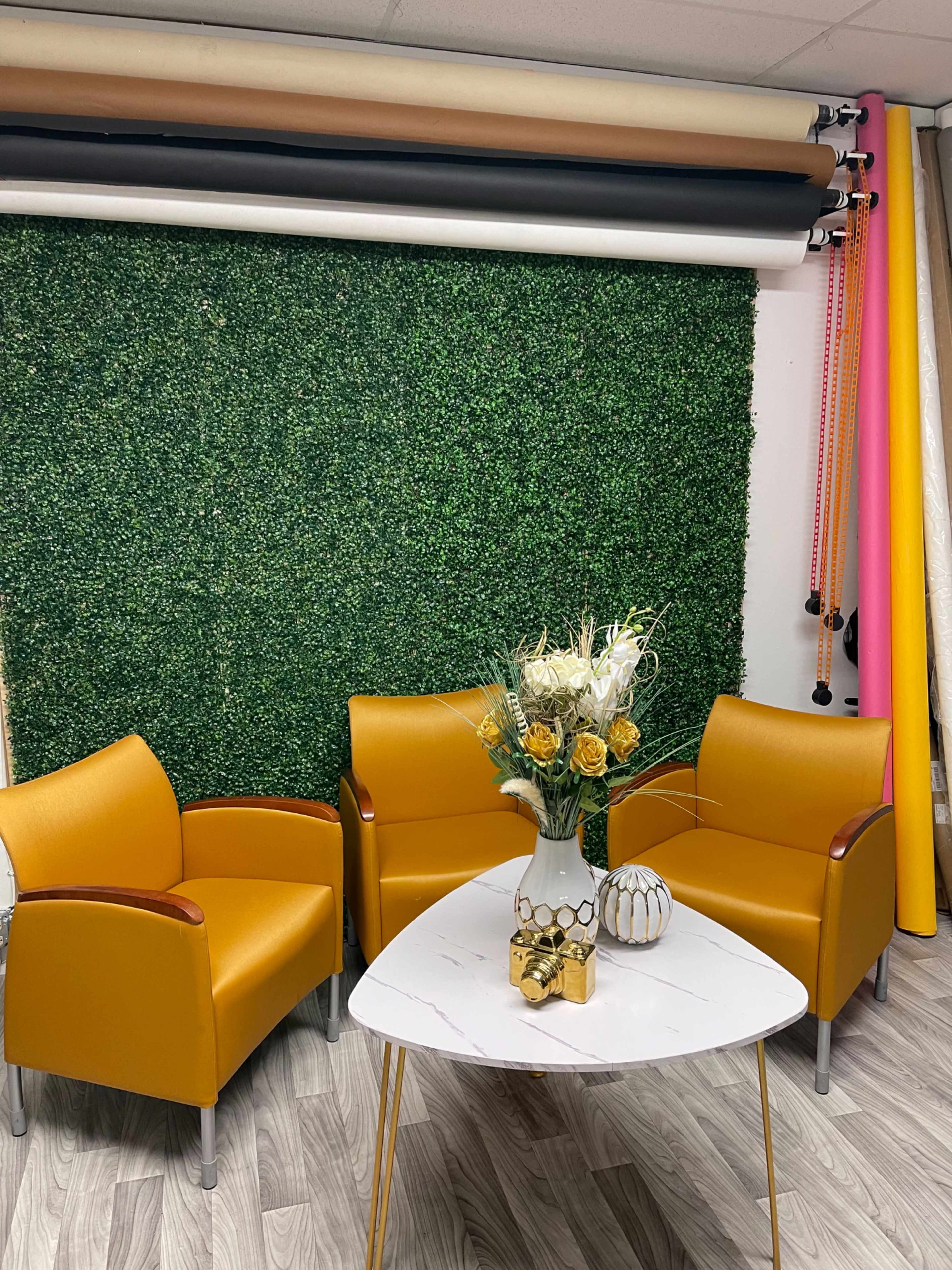 Two yellow chairs and a white table in front of a green artificial grass wall, with a flower arrangement on the table.
