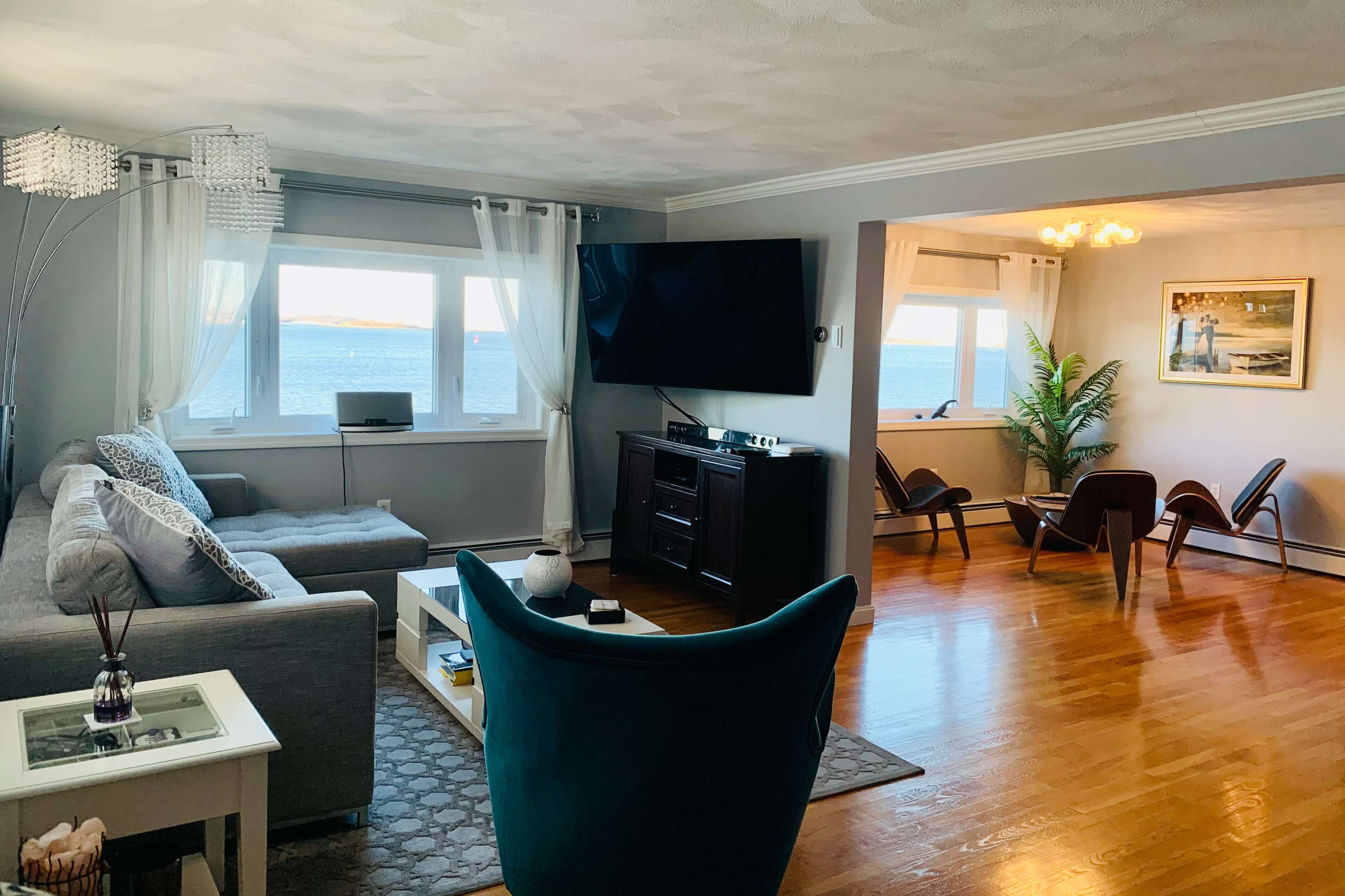 The living room features a large window overlooking the ocean, with a gray couch and a blue accent chair arranged around a coffee table and entertainment center.