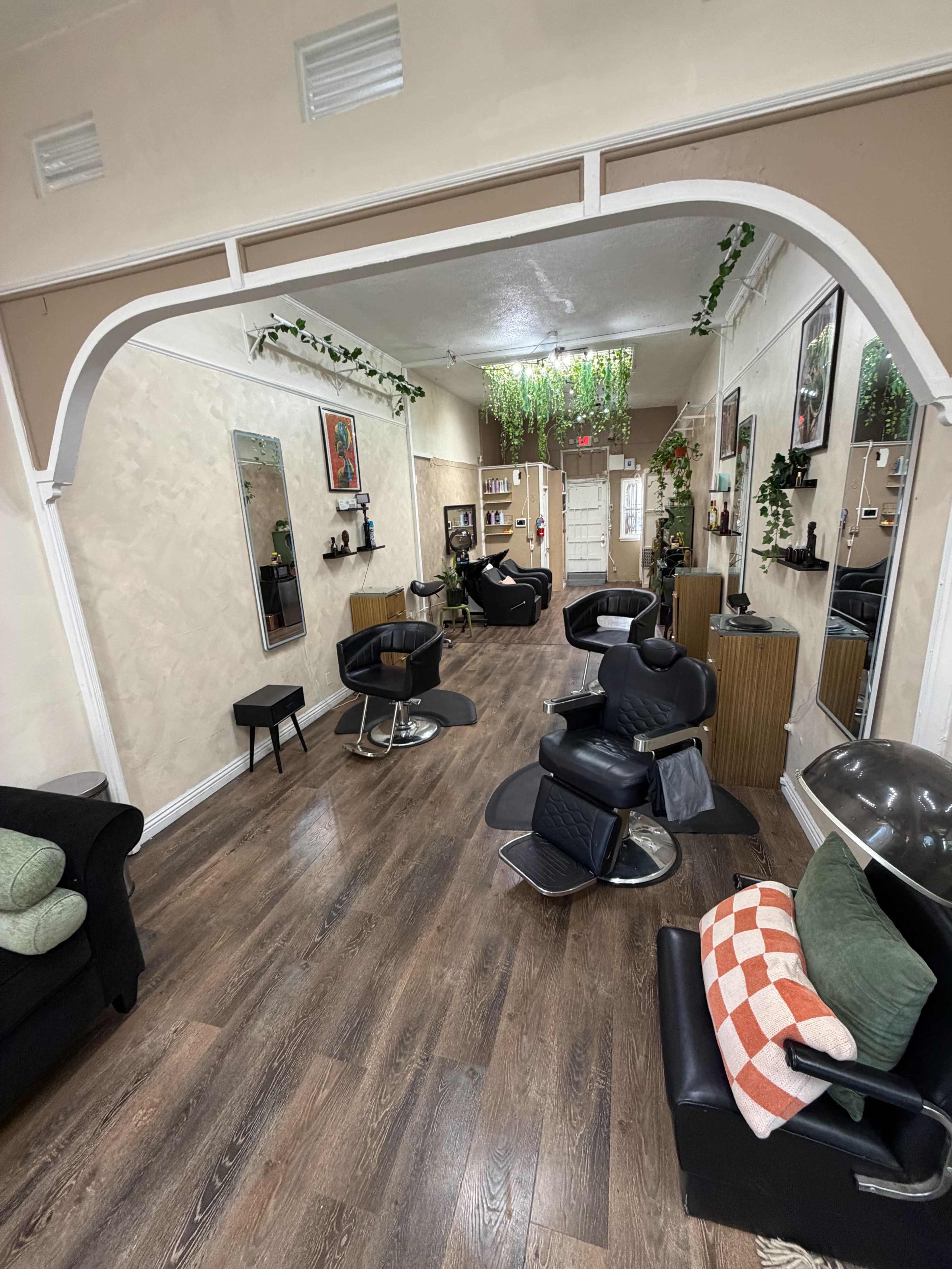 The image shows a hair salon interior featuring black styling chairs, wooden flooring, and decorative plants on the walls.