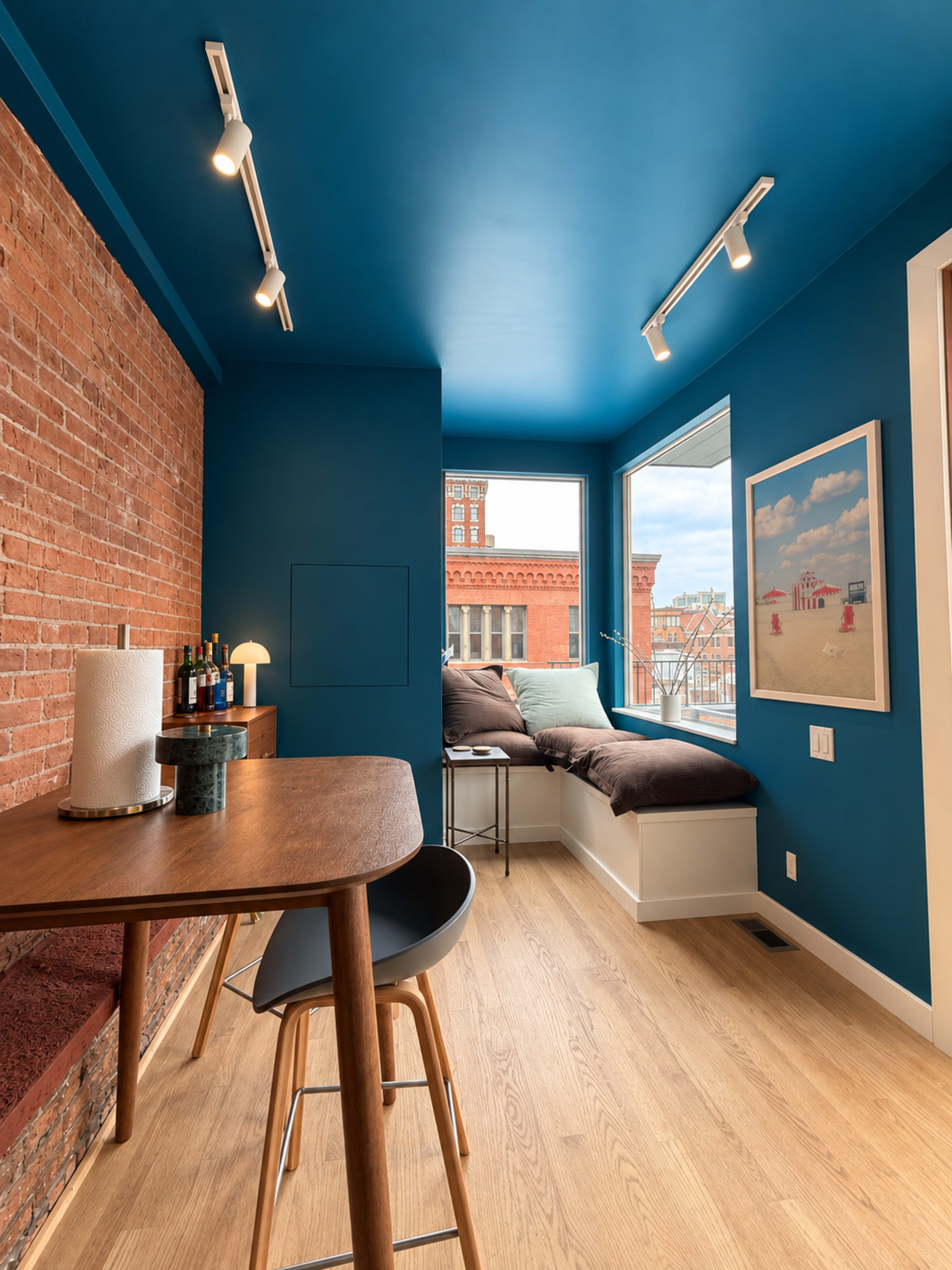 The image shows a small room with a blue ceiling, brick wall, a wooden table with a stool, and a window seat with cushions.