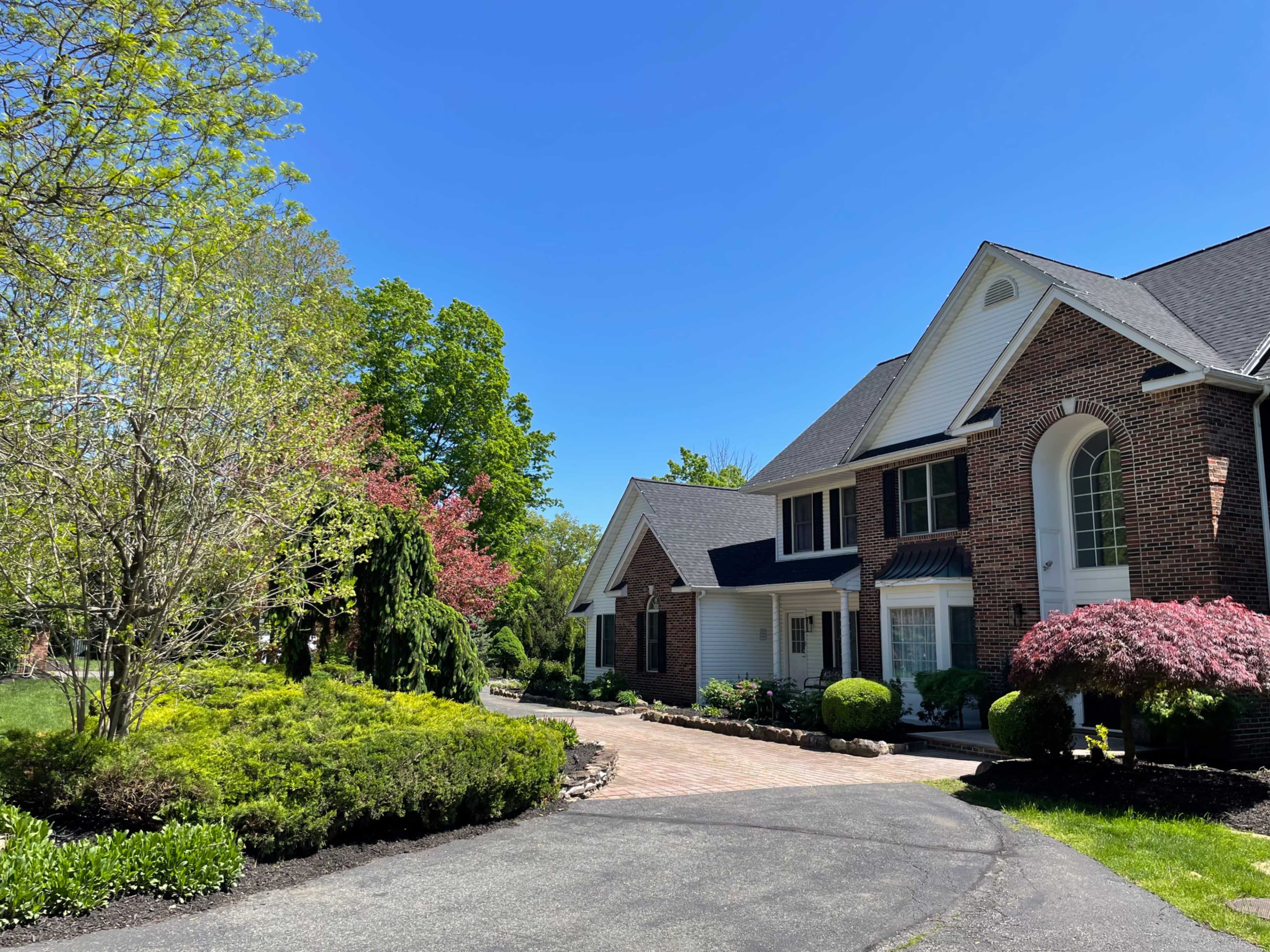 A large brick house is situated beside a landscaped driveway, surrounded by various trees and flowering plants under a clear blue sky.