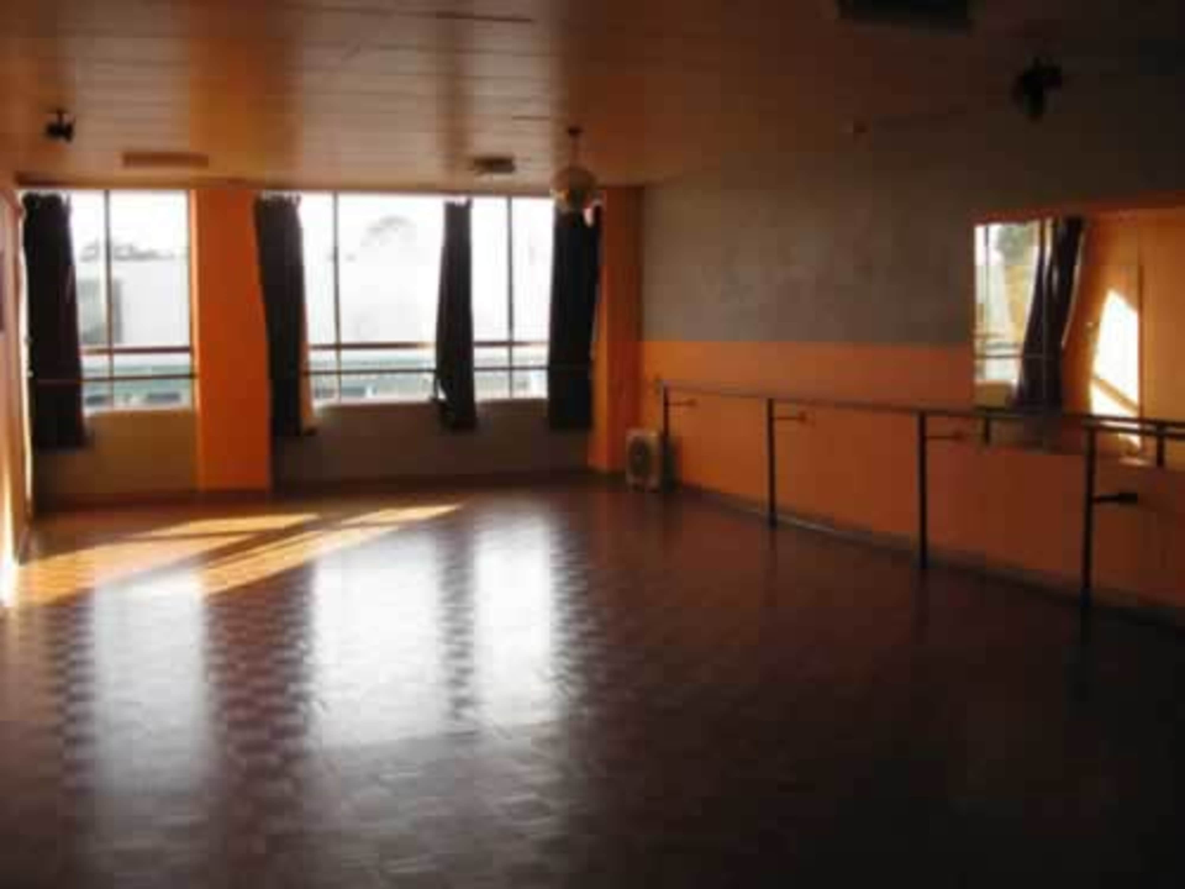 A well-lit dance studio features large windows, orange walls, and a mirrored wall along with a wooden floor and ballet barre.