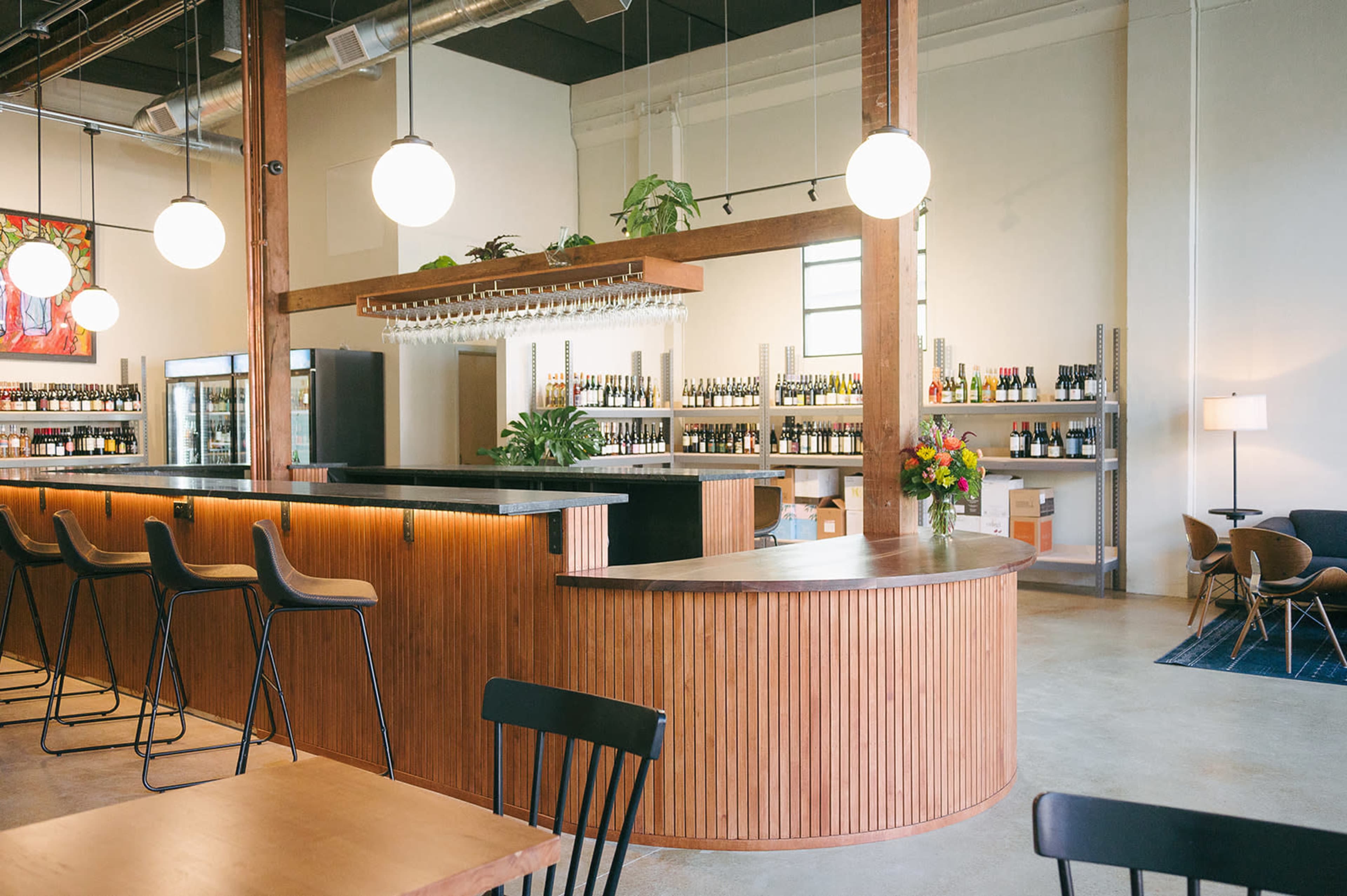 A modern bar with a curved wooden counter and high stools, surrounded by shelves stocked with various bottles in a well-lit space.