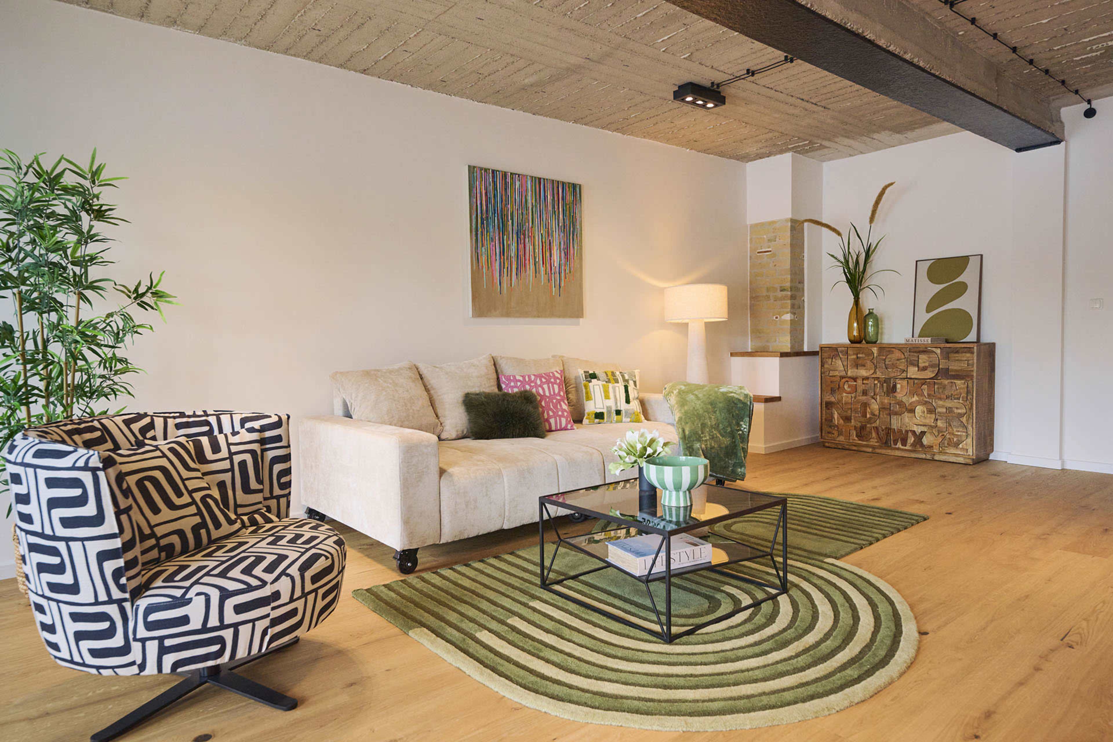 The image shows a modern living room featuring a beige sofa, a patterned armchair, a glass coffee table, and decorative plants, with light wood flooring and minimalist decor.