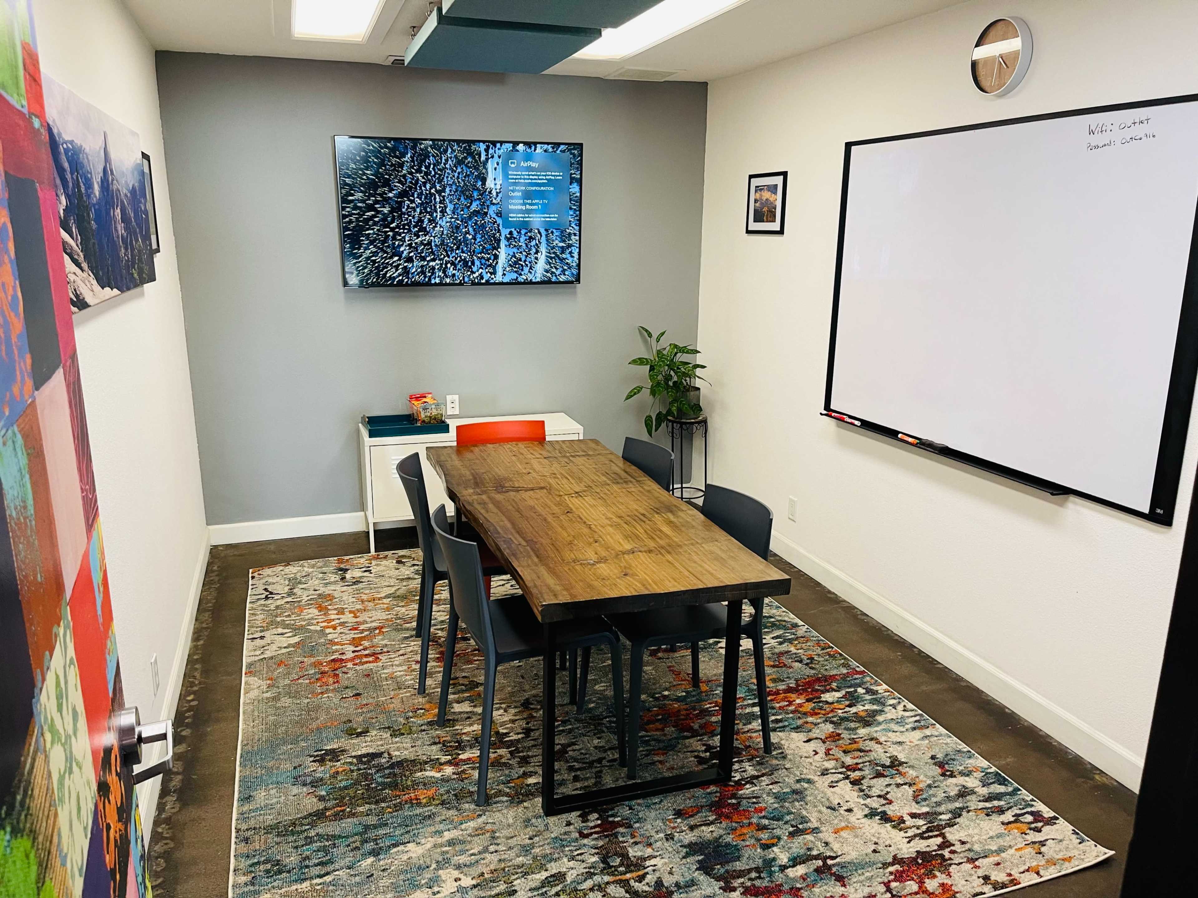 A small conference room features a wooden table surrounded by chairs, a large TV on the wall, a whiteboard, and a plant in the corner.