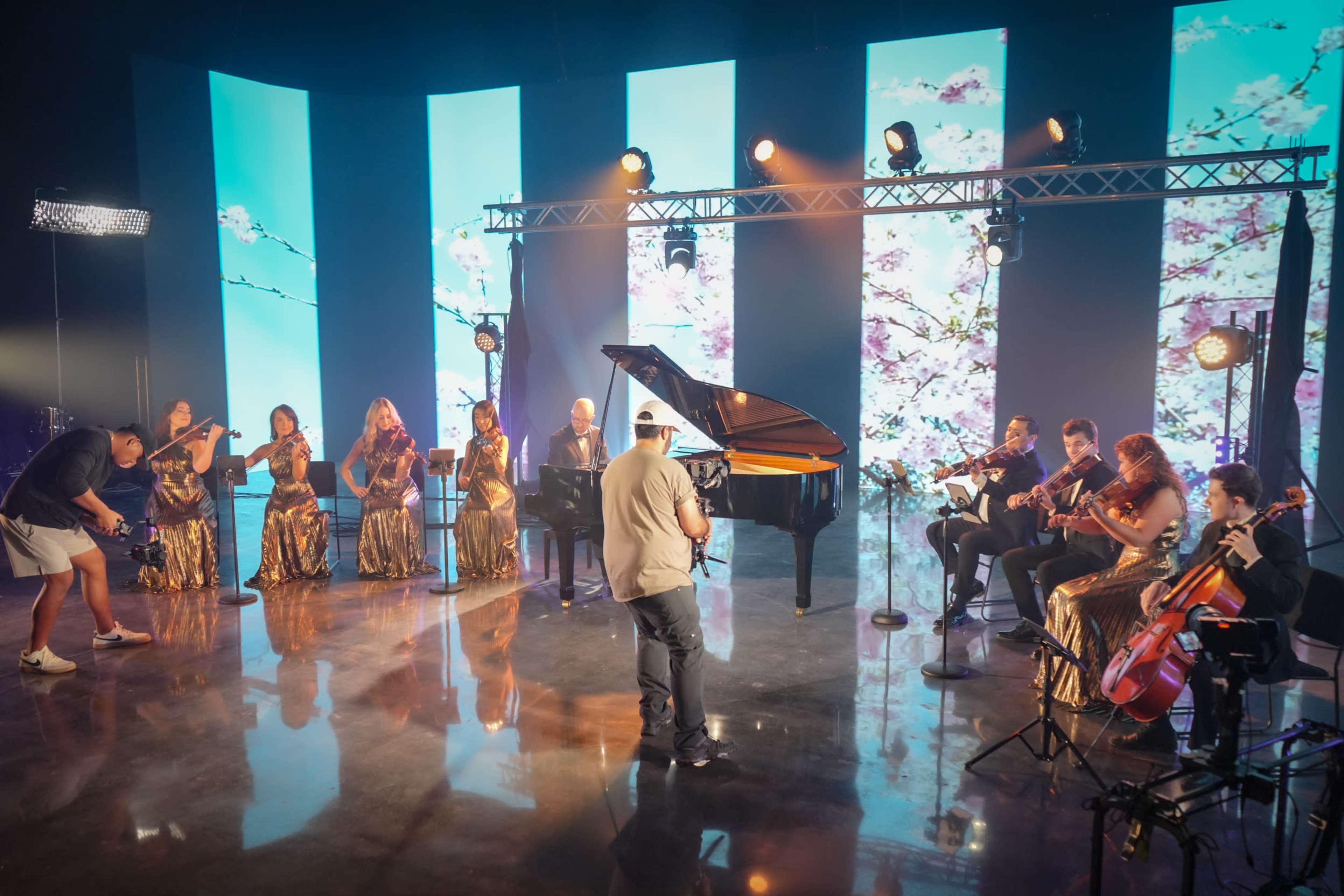 A musician performs in front of a grand piano accompanied by a string ensemble on a brightly lit stage with a floral backdrop.