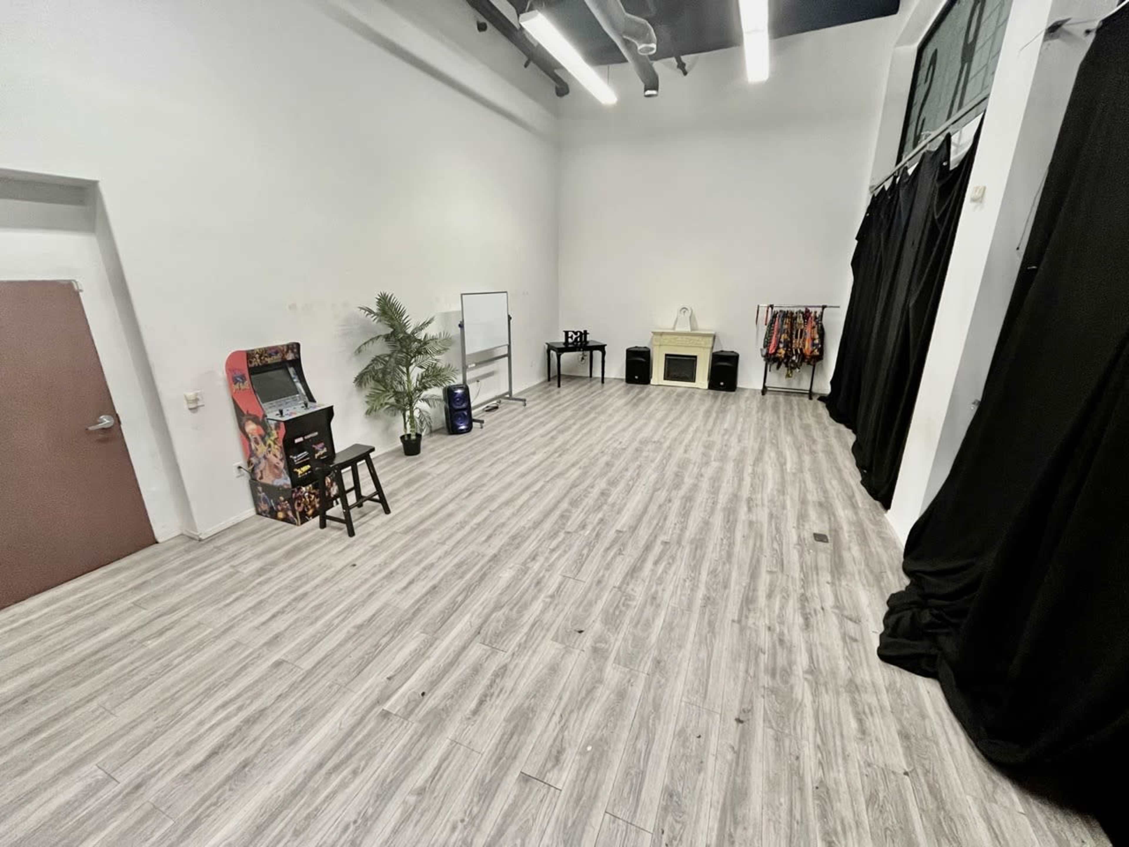 A spacious, empty indoor room with light-colored wooden flooring, a mirrored wall, and some furniture items along with a black curtain.