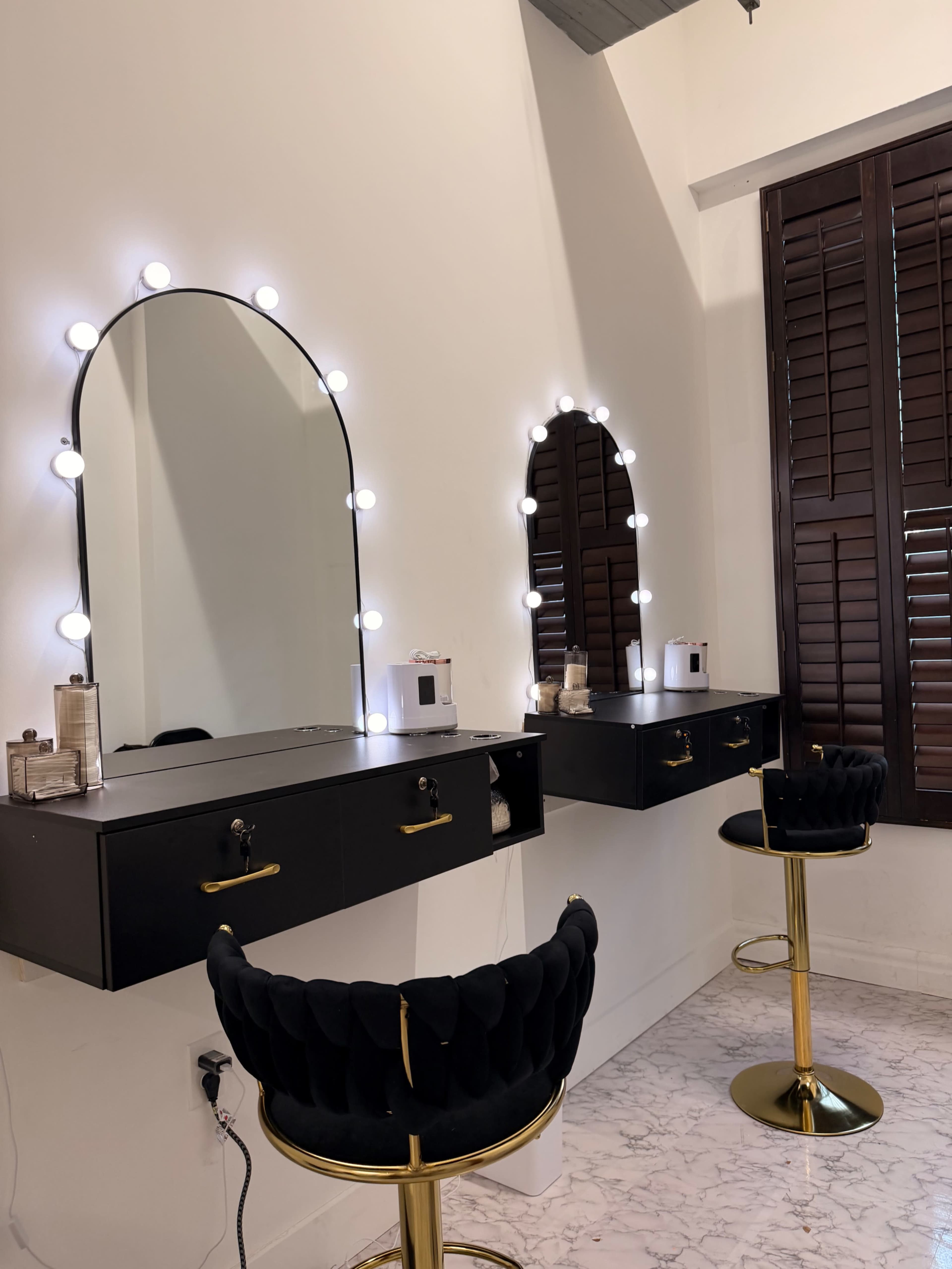 The image shows a modern makeup vanity area with two illuminated mirrors, black drawers, and stylish gold-accented stools on a marble floor.