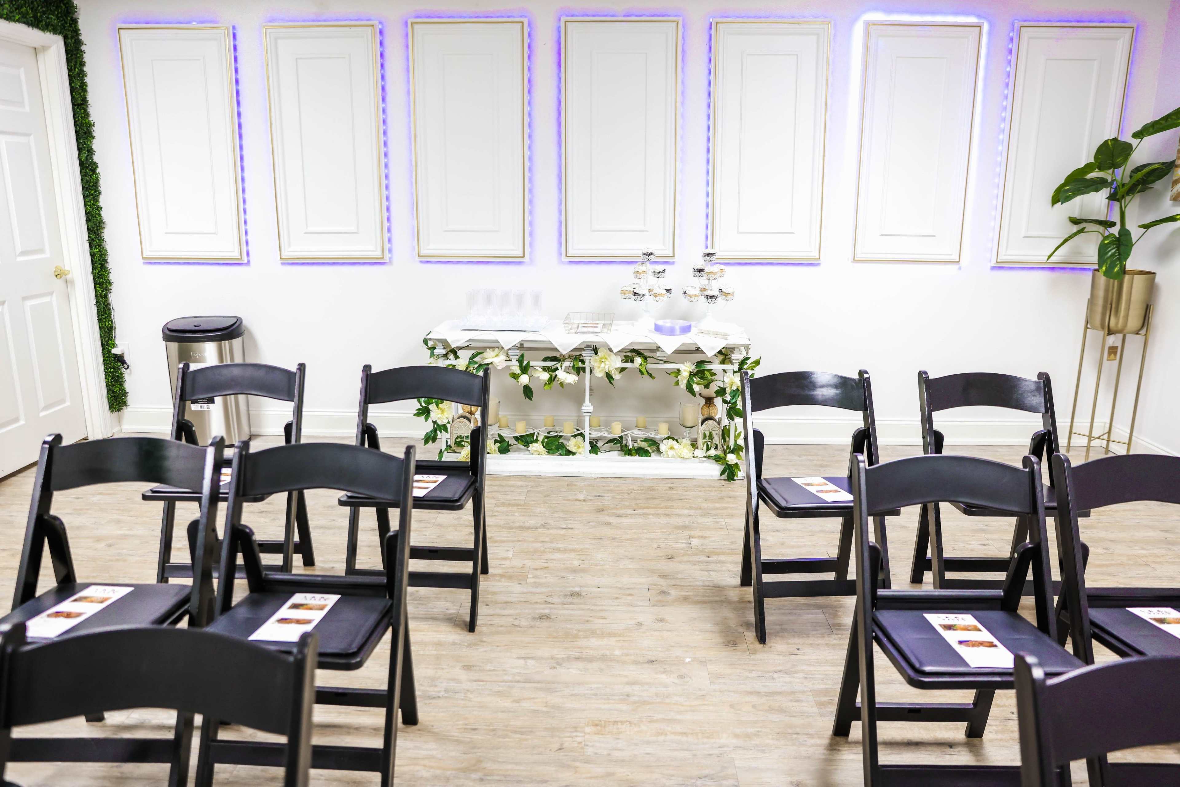 A rows of black folding chairs face a decorative table with refreshments in a room featuring white walls and framed panels.