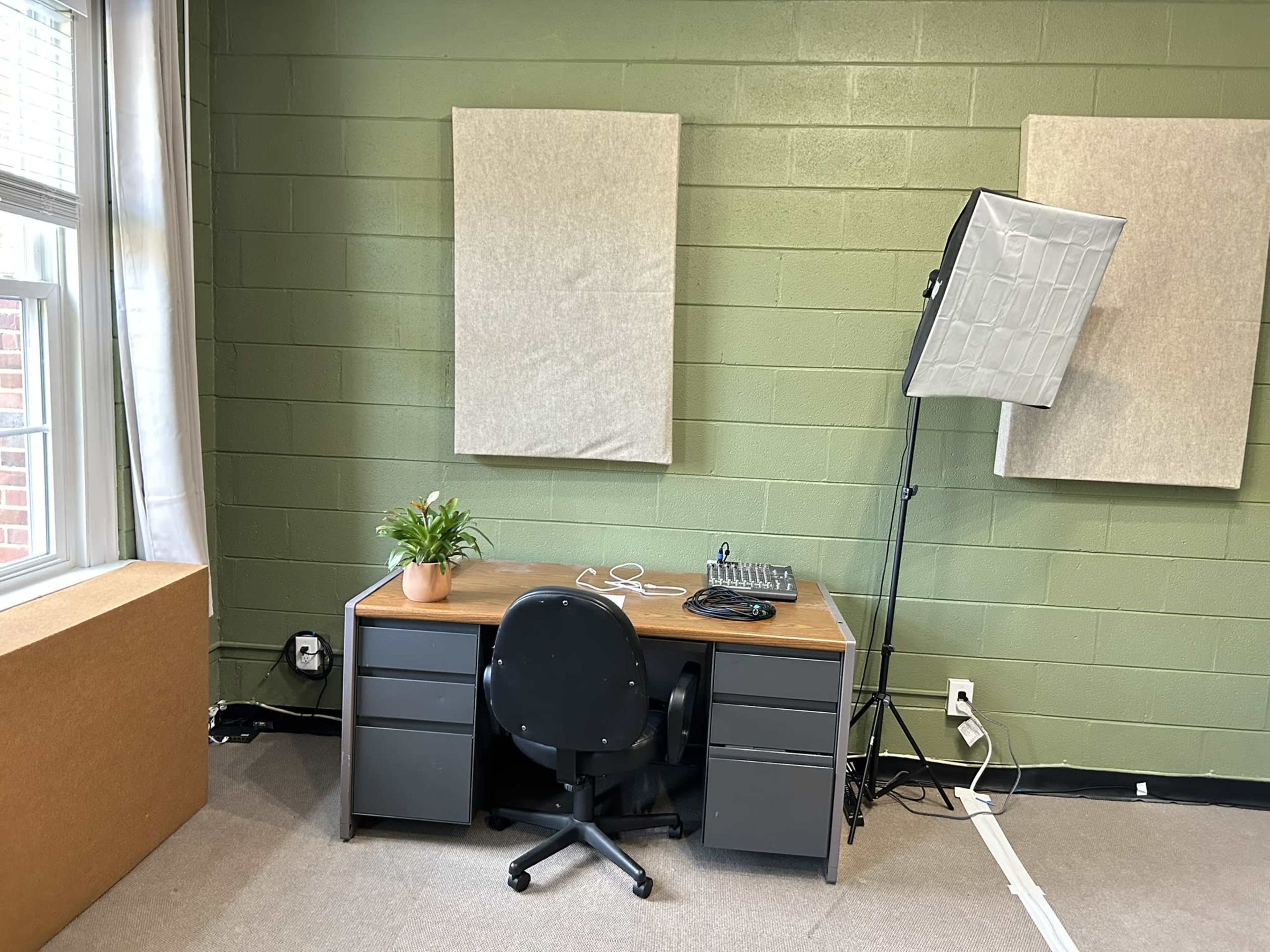 The image shows a simple office space with a desk, a black chair, a potted plant, audio equipment, and two soundproof panels on the wall.