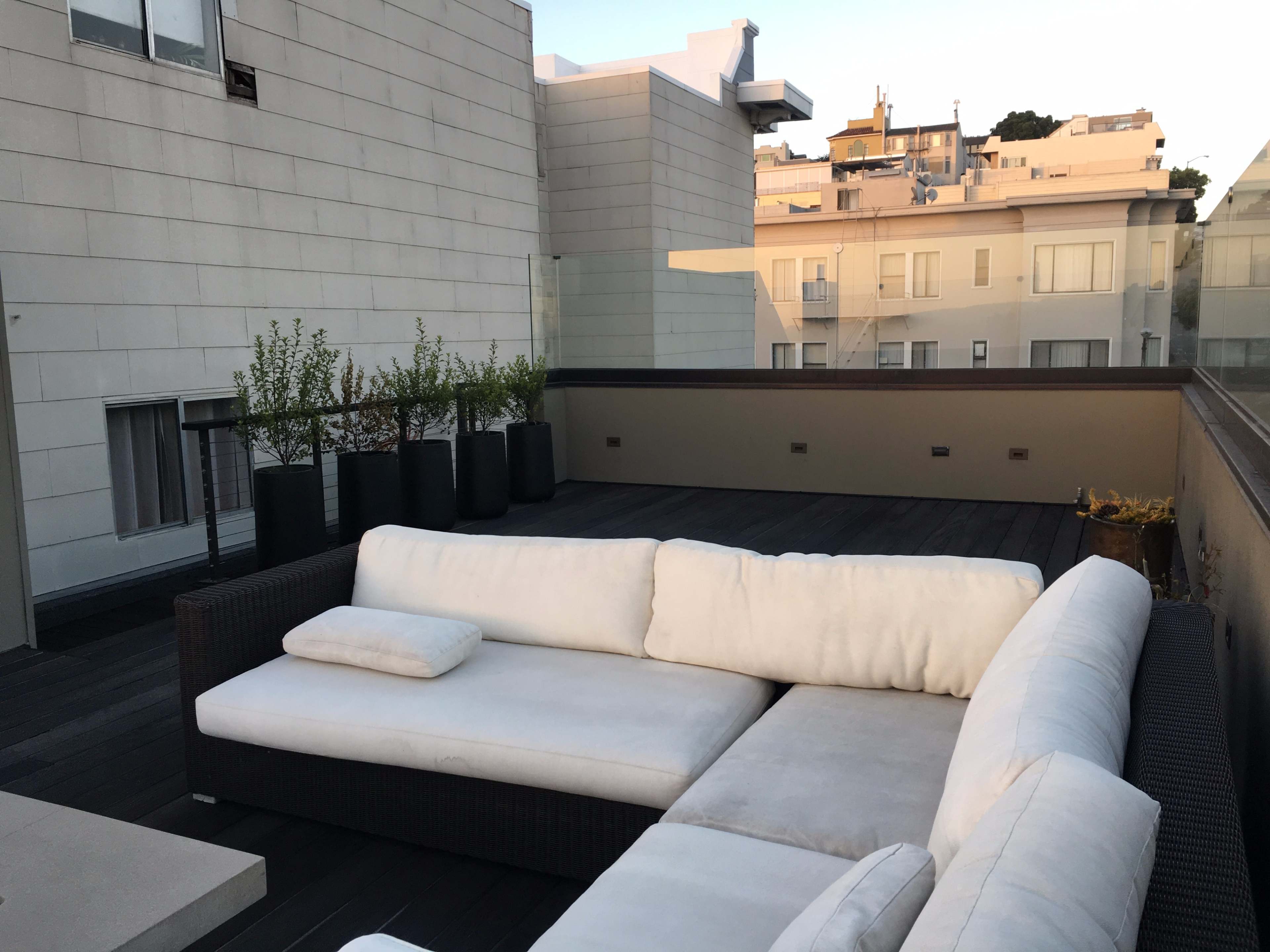 A spacious rooftop terrace features a large white sectional sofa and tall planters, overlooking city buildings.
