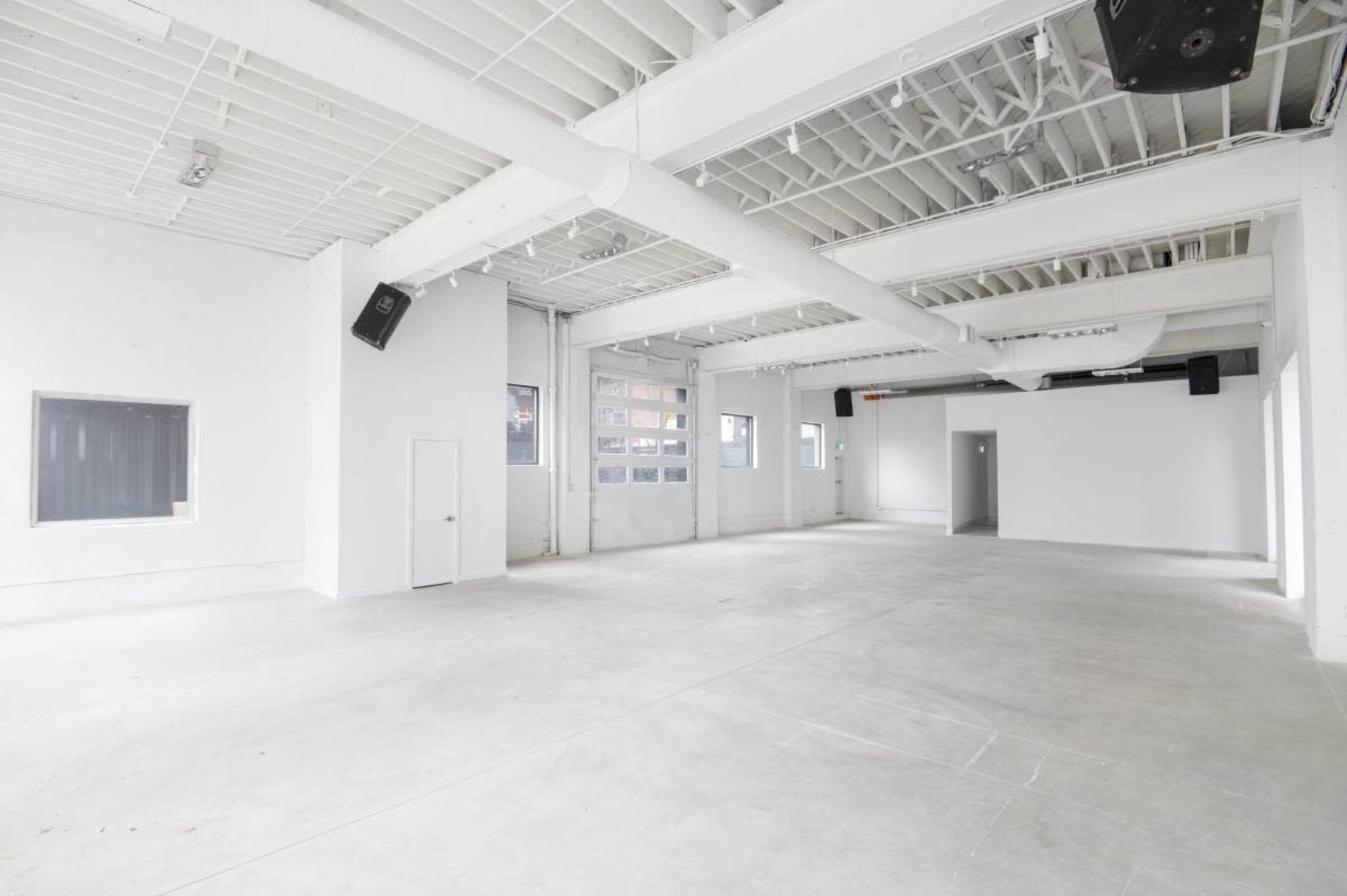 The image shows a large, empty room with white walls, high ceilings, and concrete floors, featuring multiple windows and doors.
