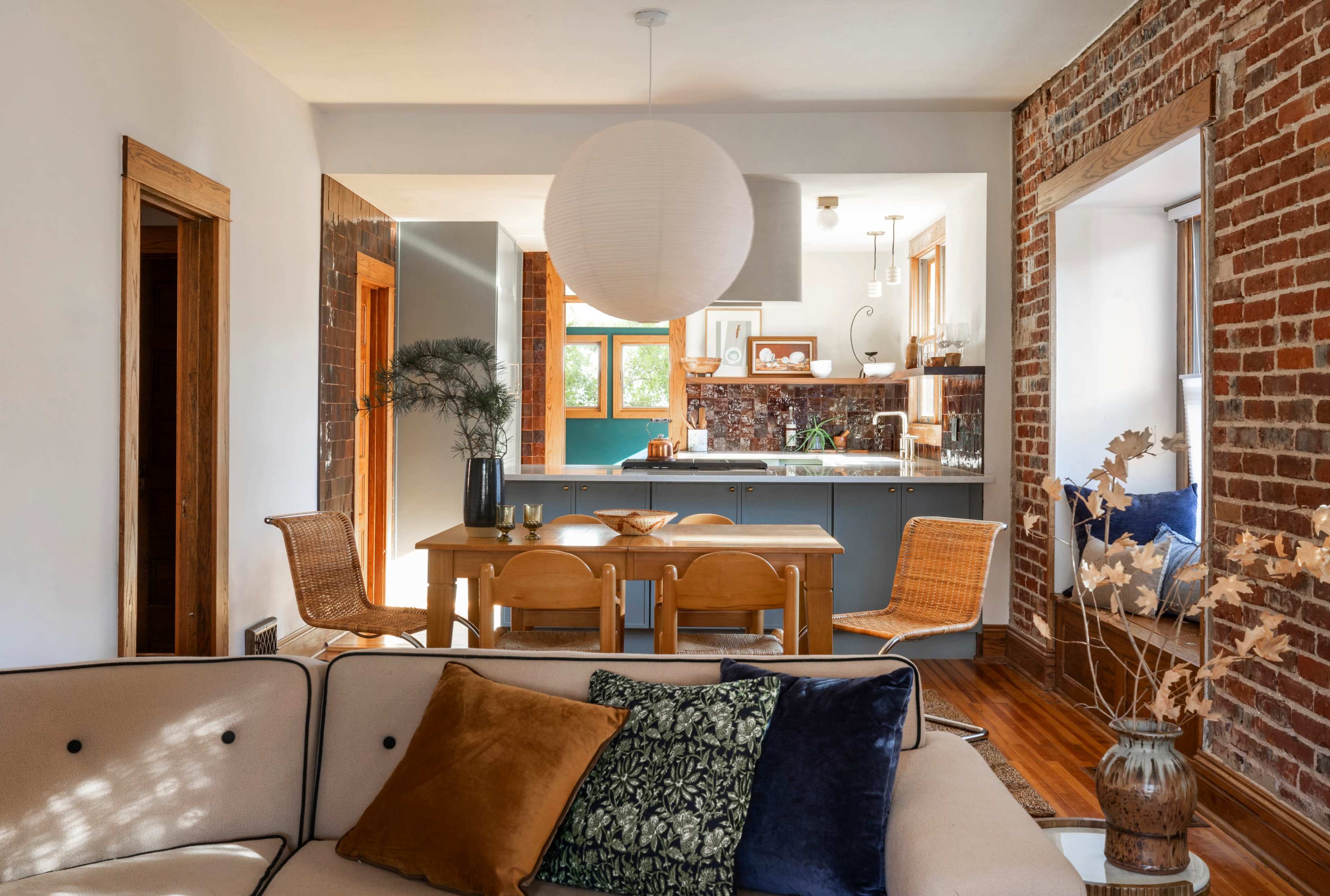A cozy living space features a sofa with cushions, a wooden dining table surrounded by chairs, and a kitchen area visible in the background with exposed brick walls.